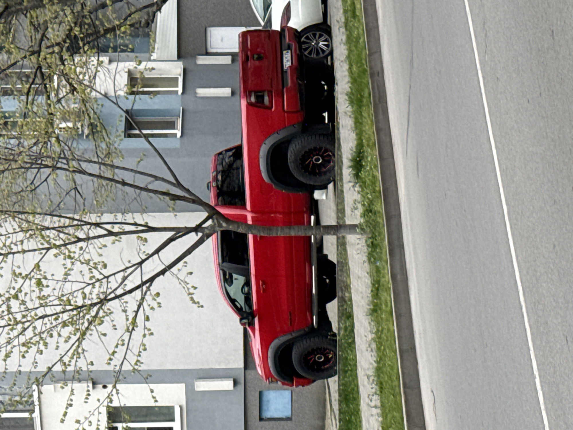 A red pickup truck is parked on the side of a road next to a sidewalk with grass, in front of a gray building with multiple windows. A tree stands between the truck and the building, its branches partially obscuring both. Another vehicle, possibly a white SUV, is parked behind the pickup truck. The location appears to be an urban area, potentially within the city of Stara Zagora, Bulgaria, based on the provided context. The road has a white line marking its edge. The photograph is taken from an elevated perspective.