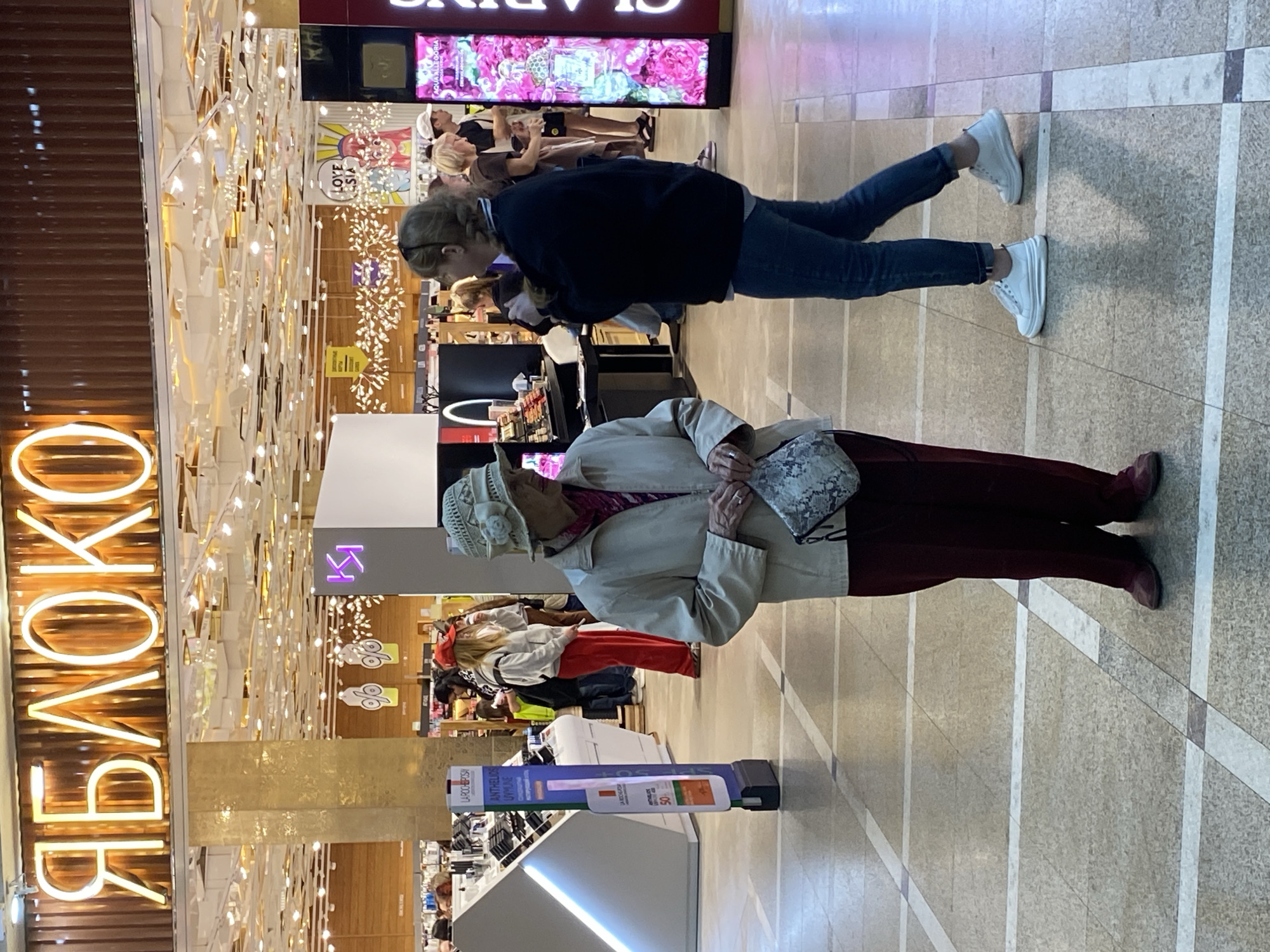 The image depicts an indoor scene within a modern retail space, likely a department store or cosmetics shop in a mall, given the provided context of Yekaterinburg, Russia. The setting is brightly lit with overhead lights and decorative light fixtures, casting reflections on the polished floor.
In the foreground, two women are visible. A younger woman, positioned towards the right, wears a dark jacket, blue jeans, and white sneakers. She appears to be looking at items on a display counter or shelf. Closer to the left and slightly behind her, an older woman stands, wearing a light-colored jacket, a patterned hat, and dark red trousers, carrying a small patterned handbag. She seems to be observing the surroundings. Several other shoppers are visible in the background, browsing various displays.
Notable details include a large, vertical neon sign on the left displaying the Cyrillic word "ЯБЛОКО" (YABLOKO), which means "APPLE" and is a well-known Russian cosmetics and perfume chain. On the right, a sign for the cosmetics brand "CLARINS" is visible above a display featuring pink floral designs and text like "AQUA ALLEGORIA" and "SOLA COCOA." Another light-up display in the mid-ground shows "LOVE ASIA." A blue and white product stand prominently features "LAROCHE-POSAY" and "ANTHELIOS UVMLINE SPF 50," indicating skincare products. The floor is tiled with a light-colored pattern and intersecting dark lines. The overall atmosphere suggests a casual shopping experience during store hours.