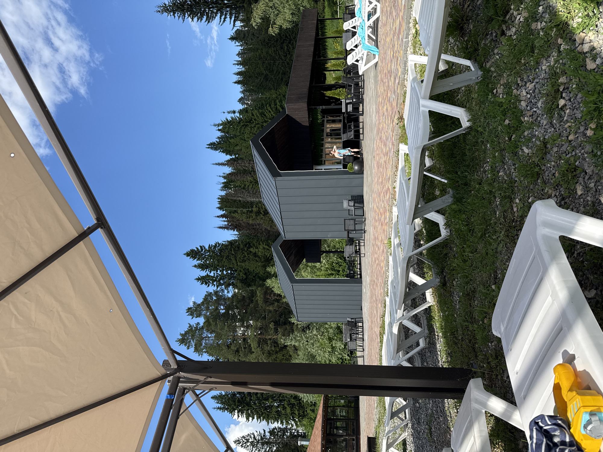 The image shows an outdoor seating area with white lounge chairs on gravel and grass, some of which are partially covered by a light-colored awning. In the background, there are modern, dark grey buildings with pitched roofs, and a forested area of tall pine trees. A person in a blue and white patterned garment is standing in front of one of the buildings. A yellow toy is visible on one of the lounge chairs in the foreground. The sky is clear and blue with a few white clouds. The scene appears to be a resort or recreational area in a natural setting.