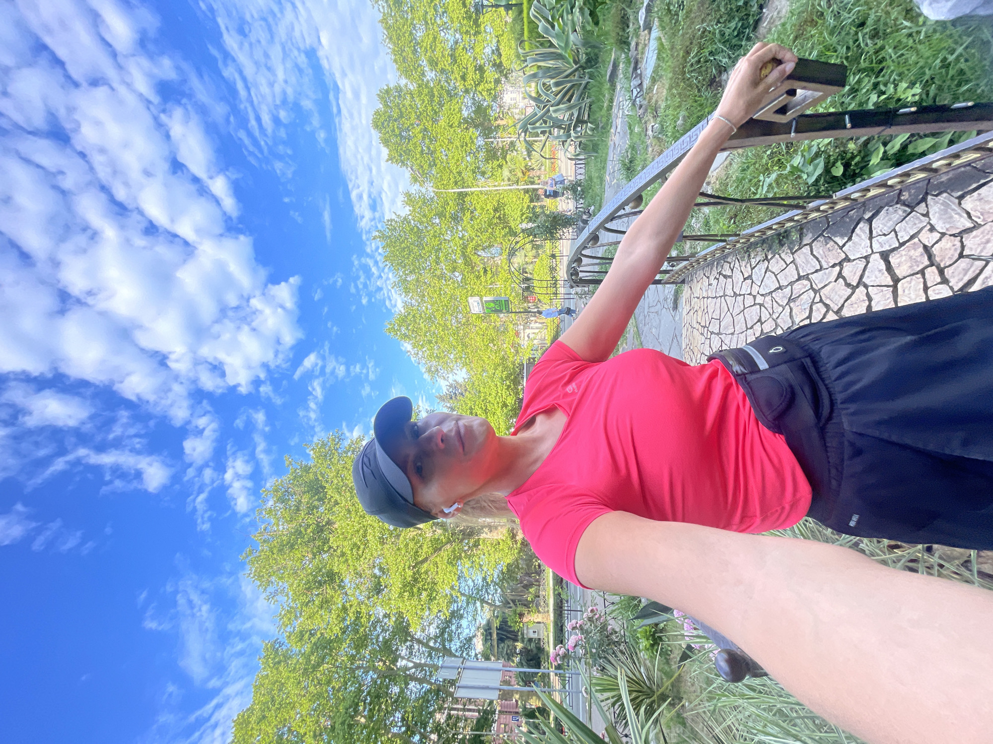 A woman is taking a selfie on a stone bridge in a park in Sochi, Russia. She is wearing a bright pink t-shirt, black athletic shorts, a black baseball cap, and white earbuds. Her left arm is extended towards the camera, and her right hand is resting on the bridge's railing. The bridge is surrounded by lush greenery, including trees, bushes, and large plants. In the background, there are more trees, a few people walking, and some buildings. The sky is blue with scattered white clouds. The sun appears to be shining, casting light and shadows on the scene.