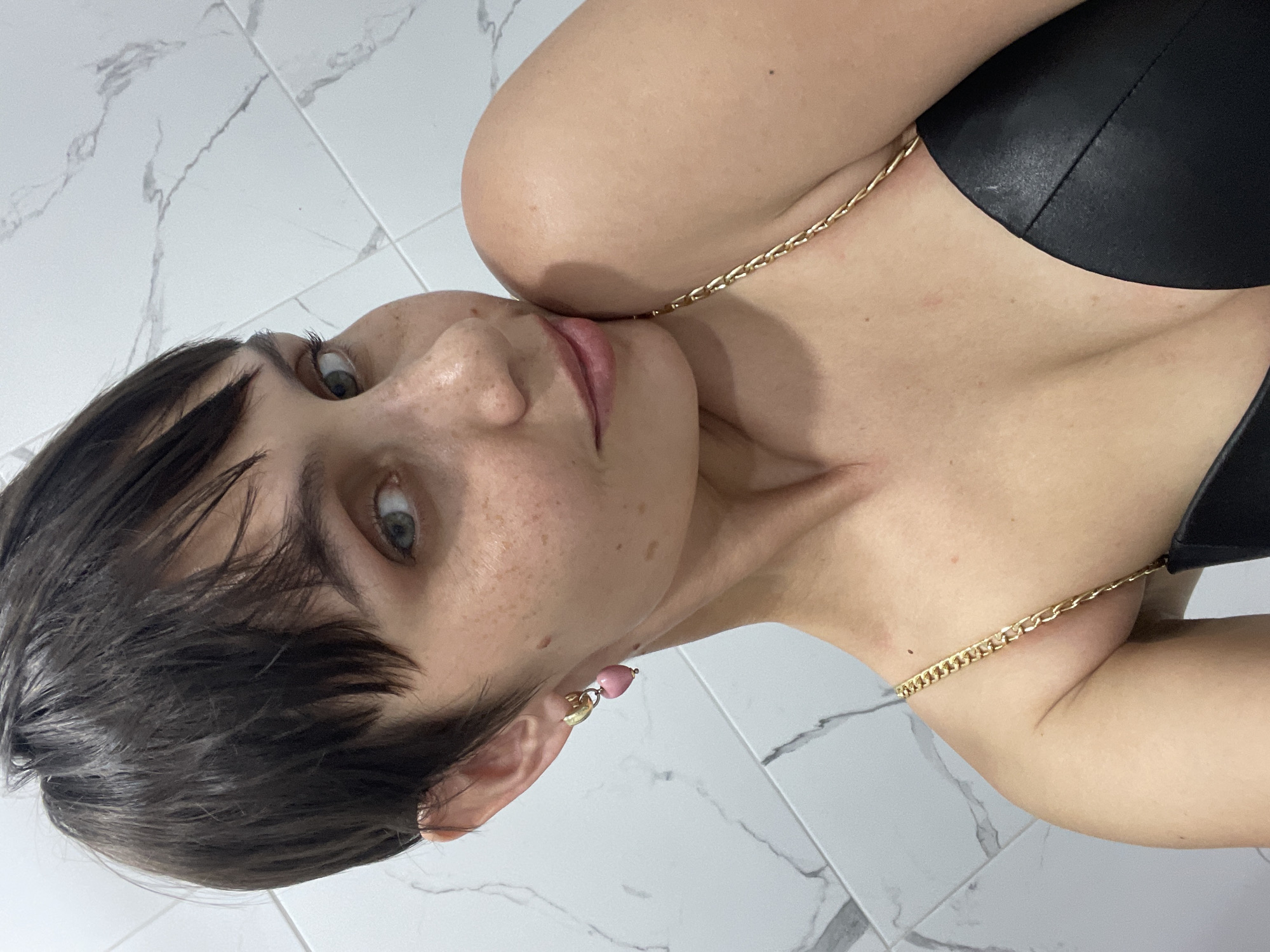 A close-up, tilted image of a fair-skinned individual with short, dark brown hair and light eyes with freckles. They are wearing a black top and a gold chain necklace. A pink heart earring is visible on their left ear. The background consists of light grey and white marble-patterned tiles. The location in the city of Izhevsk, Russia is unknown.