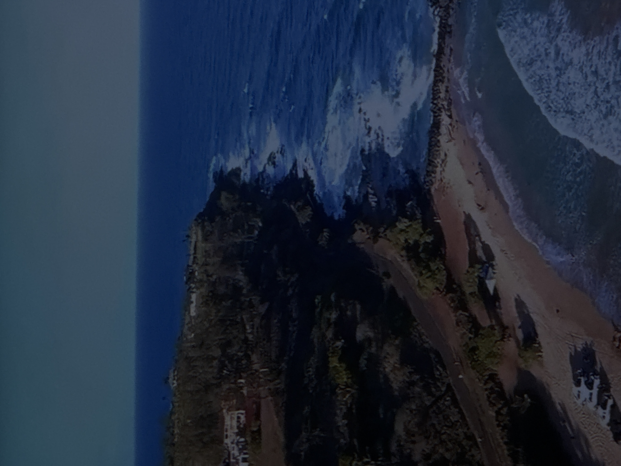 This is an aerial, oblique view of a coastal landscape. The image shows a rugged, vegetated coastline meeting a dark blue ocean with white waves. Along the cliff edge, several buildings are visible, suggesting a settlement. Further down, a sandy beach is present, with a road running parallel to it. On the beach, a few vehicles, including what appears to be a white SUV or similar vehicle, are parked. There are also some structures that could be tents or temporary shelters. The sky is a clear, light blue, transitioning to a darker blue towards the horizon. The overall scene depicts a somewhat secluded coastal area with signs of human activity. The image is not set in Sinyavino, Russia; Sinyavino is an inland town.