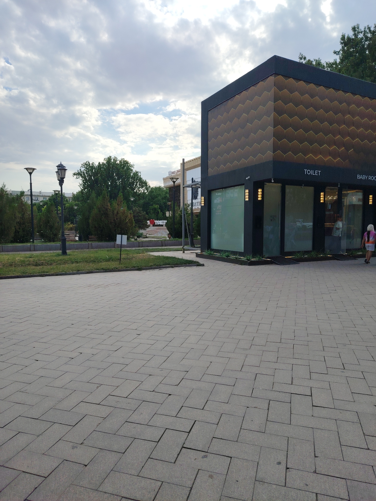 The image shows a modern public toilet building with a honeycomb-patterned facade in Shymkent, Kazakhstan. The building is located in a paved public area with a park in the background. Several streetlights and trees are visible. A young girl in a pink and white outfit is walking away from the toilet building towards the right side of the image. The sky is overcast. The ground is paved with gray rectangular tiles laid in a herringbone pattern. The toilet building has large glass windows and signage indicating "TOILET" and "BABY ROOM".