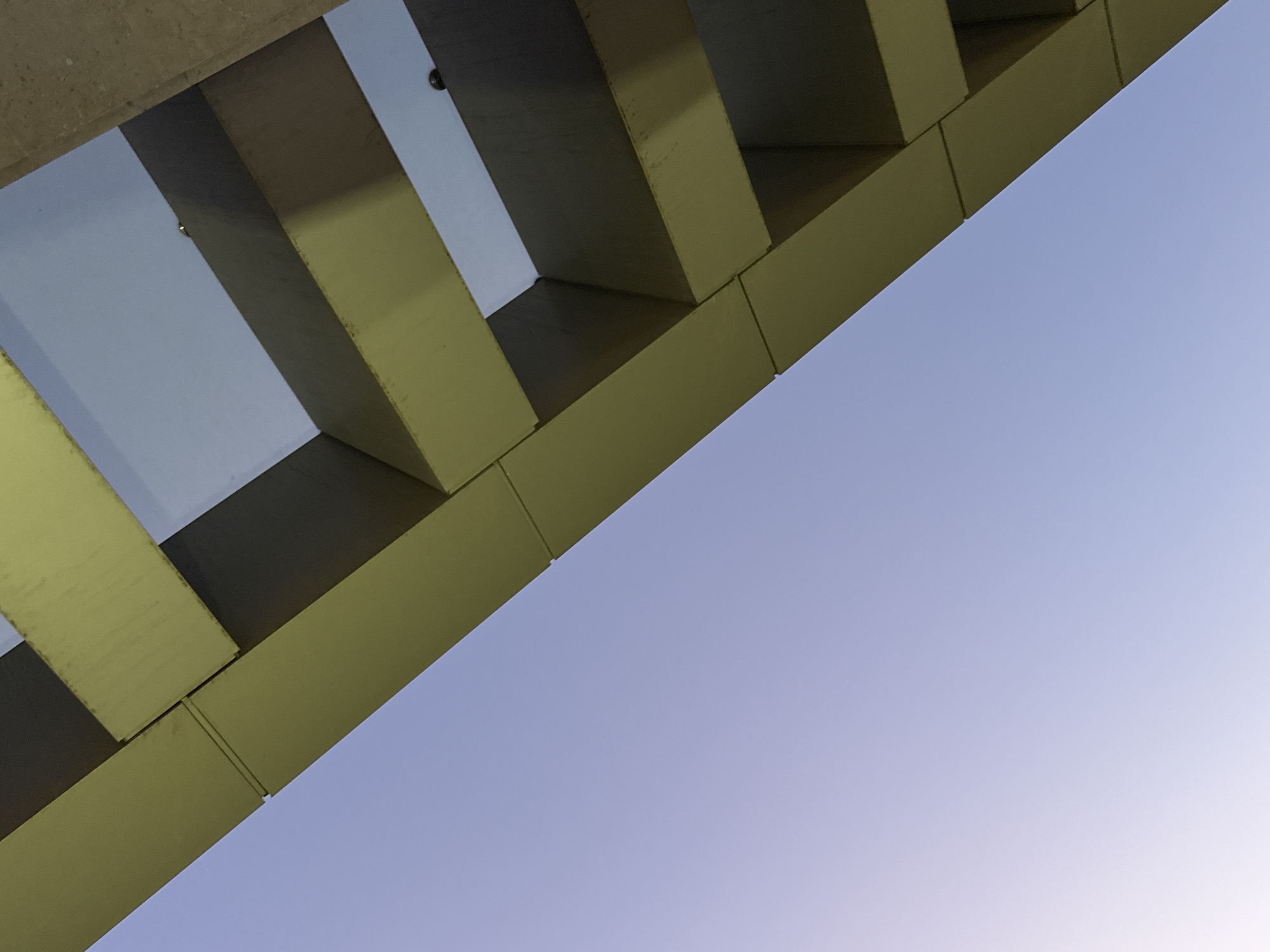 The image captures an abstract, upward-looking view of a modern architectural structure set against a clear, gradient sky. The primary subjects are a series of parallel and intersecting structural beams or girders, made of a light olive green or khaki-colored material, possibly painted concrete or metal. These beams are arranged diagonally, forming a geometric pattern across the upper left and central portions of the frame.
The architectural elements showcase clean lines and rectangular forms, with some beams casting subtle shadows on adjacent surfaces, creating a sense of depth. Visible seams and panel joints on the beam surfaces suggest a constructed finish. Faint, dark, circular objects, possibly embedded fixtures or bolts, can be discerned within the gaps between some of the beams. A small section of a rougher, lighter grey surface, possibly raw concrete, is visible in the extreme top-left corner.
The sky, which occupies the majority of the image, displays a soft gradient of purplish-blue, lightening towards the bottom right. This gentle coloration and lack of harsh shadows suggest the time of day is either early morning (dawn) or late afternoon/early evening (dusk). The weather is clear and cloudless. There are no people or discernible activities taking place, and no visible text in the image. The scene is an abstract architectural detail, located in Yerevan, Armenia.