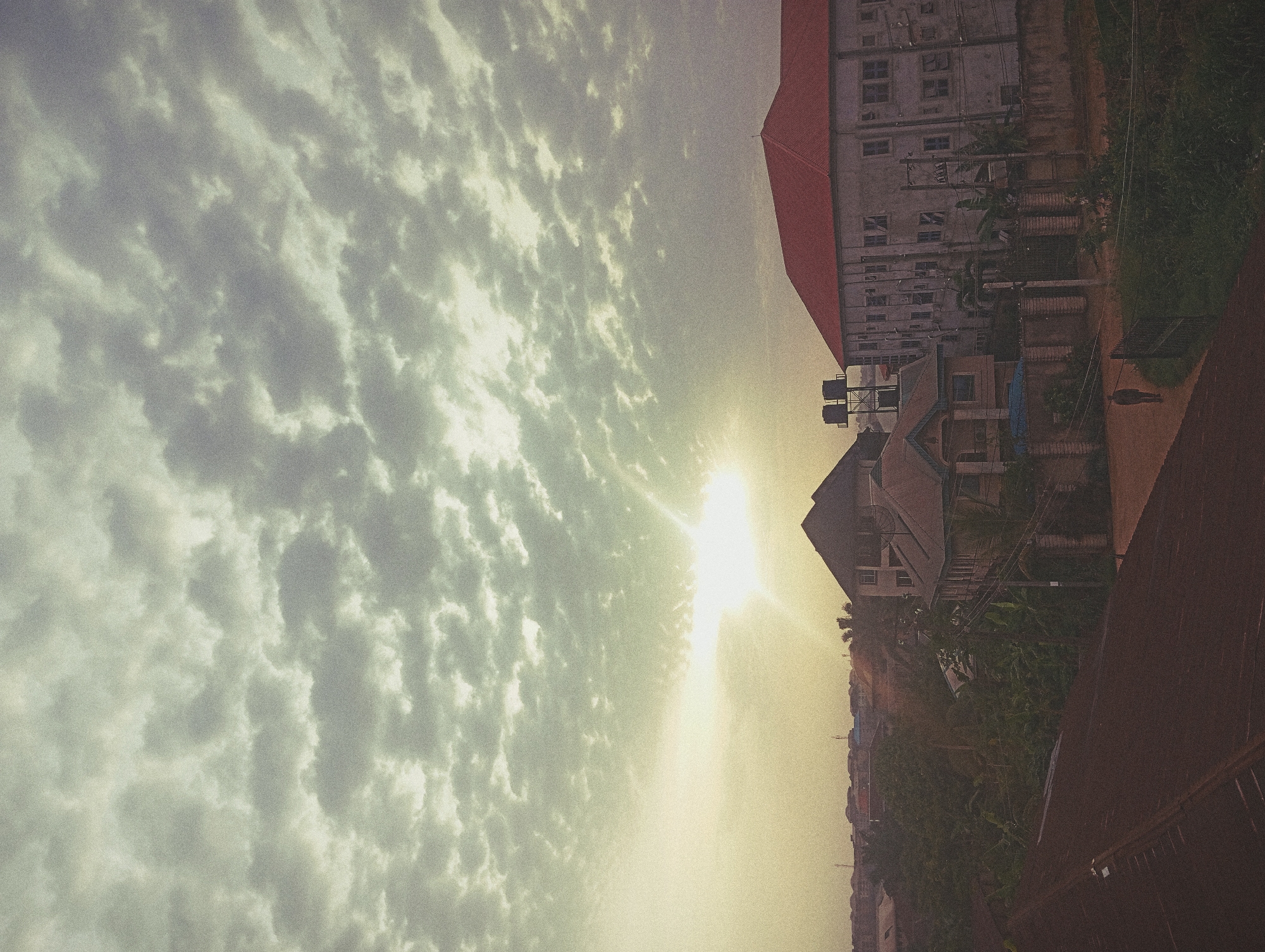 A high-angle, rotated outdoor photograph shows a sky filled with altocumulus clouds illuminated by the sun in the Nigerian city of Nkpor. Buildings are visible on the right side of the image, with a tall apartment building with a red roof directly above a small house and green vegetation. Power lines run near the tall building. At the bottom right is a partial view of a corrugated iron roof. At least one person is visible walking along a paved area near the tall building.
