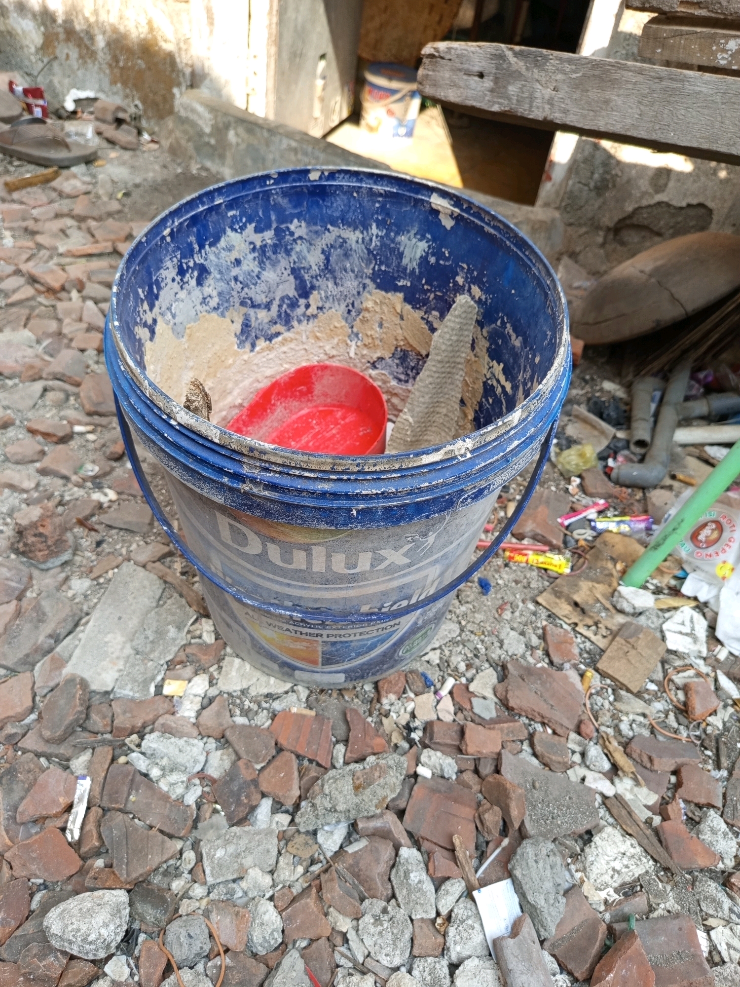 A blue plastic bucket, labeled "Dulux," is positioned on a ground covered with broken bricks and debris. The bucket contains a red plastic scoop and a partially hardened substance, possibly dried paint or plaster. The surrounding environment appears to be an outdoor area, with a concrete wall and a wooden beam visible in the background. The scene suggests an area undergoing construction or demolition, or perhaps a neglected storage space. The location is Tebel, Indonesia.
