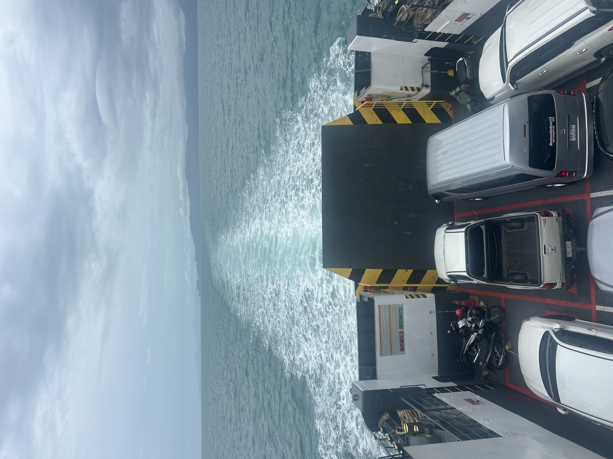 The image depicts the rear deck of a ferry as it moves through the ocean, leaving a white wake behind it. The sky above is overcast with a heavy cloud cover. In the distance, a faint, hazy outline of mountains or land is visible on the horizon.
On the ferry deck, several vehicles are parked. Closest to the viewer is a white sedan. To its right is a white pickup truck. Further back, a silver van is parked, and to the right of the van is another white sedan. In the far left of the deck, two motorcycles are parked side-by-side. A red traffic cone is visible near the motorcycles.
The ferry deck itself has a utilitarian appearance, with yellow and black striped barriers and white-painted bulkheads. A sign with text and various icons is affixed to one of the bulkheads. The overall scene suggests transportation of vehicles and people across a body of water under cloudy conditions. The location is identified as Unknown, Thailand.