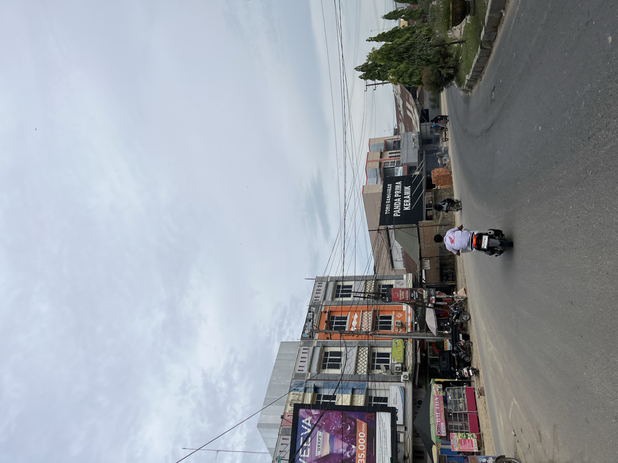 This image captures a street scene in Bundar, Indonesia, on an overcast day. The perspective is a low-angle shot taken from a street level, looking slightly upwards. The foreground is dominated by a paved road, with a single motorcycle rider in the center, wearing a white shirt with red accents and dark pants. They are facing away from the camera, moving along the road.
To the right of the road, a row of multi-story buildings lines the street. The buildings exhibit a mix of architectural styles and colors, with some featuring modern facades and others showing signs of age. One building prominently displays a black banner with white text that reads "TOKO BANGUNAN PANDA PRIMA KERAMIK." Another building, painted in vibrant orange and gray, has multiple windows and air conditioning units. Below these buildings, several small shops and kiosks are visible, some with colorful awnings. A large billboard advertising "GRAPE" and "35.000/POD" is partially visible at the bottom left.
On the left side of the road, a grassy embankment slopes upwards, with trees and power lines creating a slightly cluttered urban landscape. Several other motorcycles are parked or in motion further down the street, indicating a typical day in a populated area. The sky is cloudy, suggesting either early morning or late afternoon, or simply a day with overcast weather. The overall impression is one of a busy, functional street in an Indonesian town.