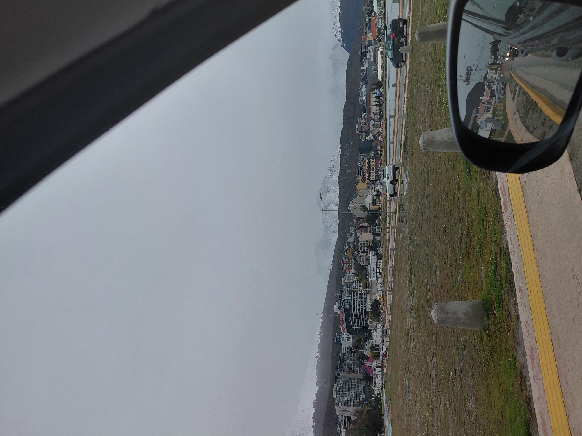 The image depicts a scene in Ushuaia, Argentina, viewed from inside a vehicle. The city, situated along the Beagle Channel and backed by snow-capped mountains, is visible under a cloudy sky. The landscape consists of a mix of buildings and vegetation. On the right side of the frame, a side mirror shows a road with cars. The side of the road has grass, concrete posts, and a sidewalk with a yellow tactile paving surface. The foreground includes part of the interior of the car, including the window frame.