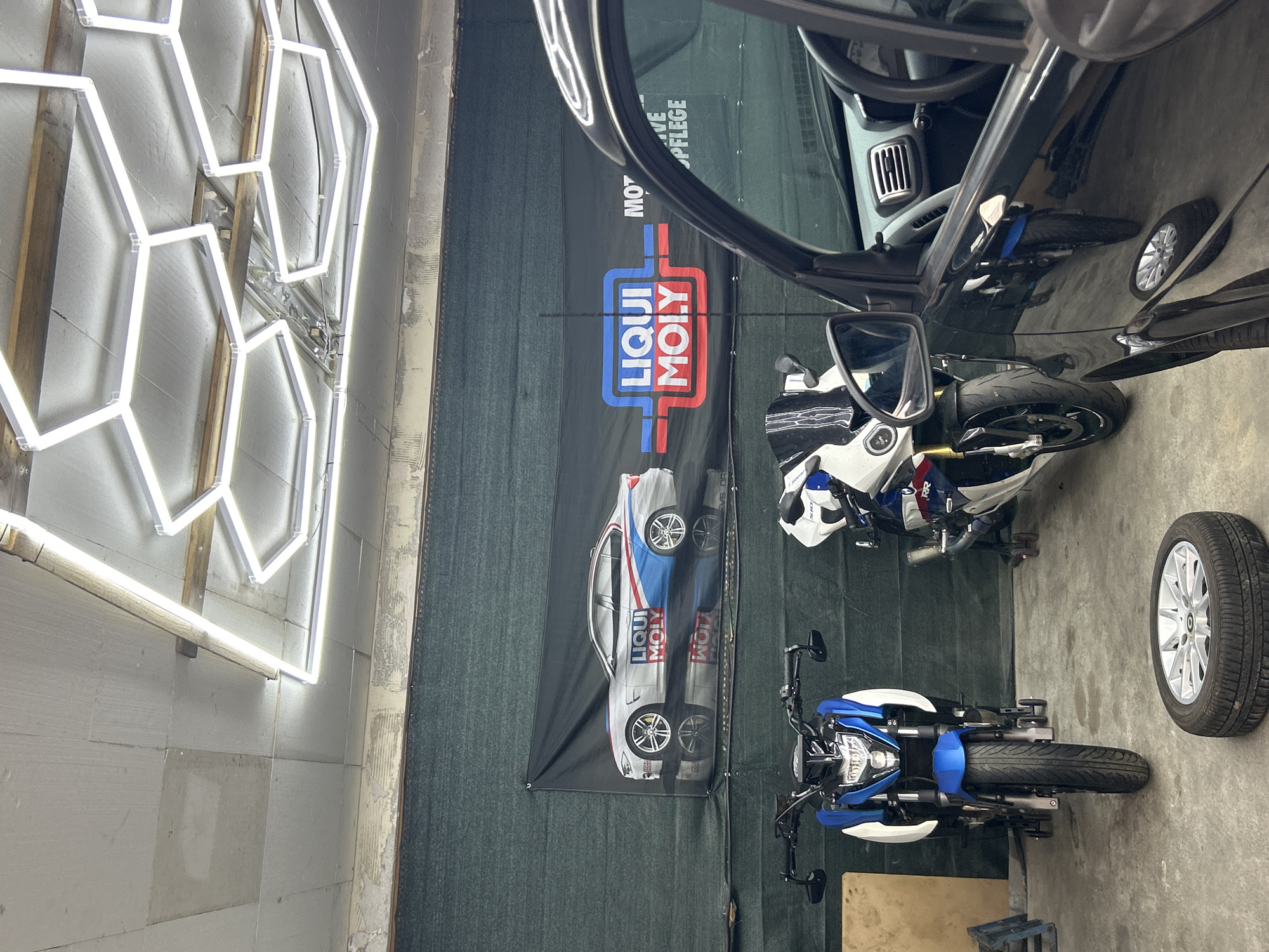 In an indoor setting, possibly a garage or workshop, a black car is parked to the right of the frame, partially obscuring two motorcycles and a banner. The banner, displayed on the back wall, features a silver car with "LIQUI MOLY" branding. In front of the banner, a blue and white motorcycle stands on the left, and a white, blue, and red motorcycle is on the right. A tire and wheel sit on the concrete floor in the foreground. Above, a modern hexagonal light fixture illuminates the space, attached to a wooden ceiling structure. The environment appears utilitarian, with exposed ceiling elements and a dark green tarp or wall covering. There are no people visible in the image.