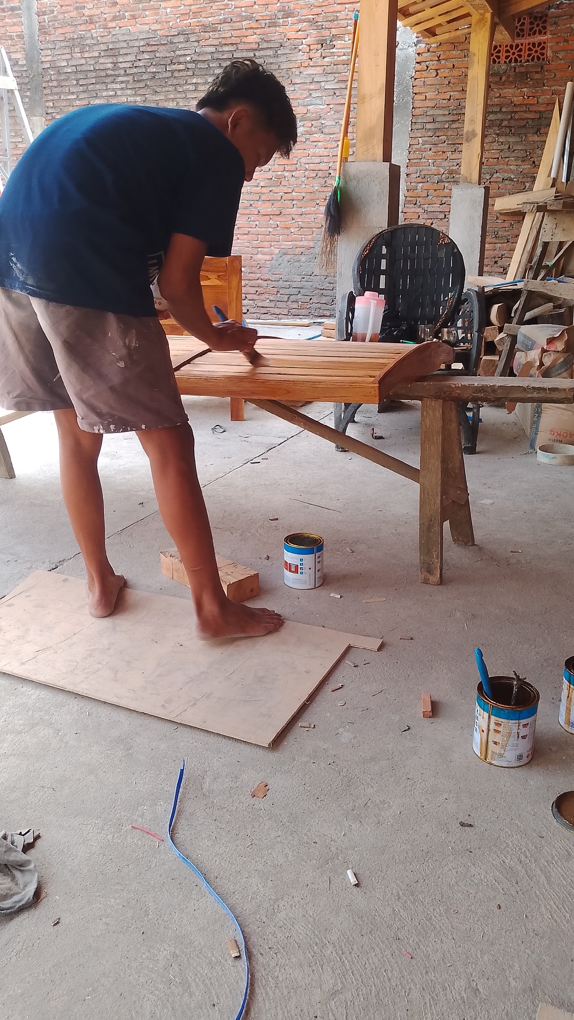 In Karanglangu, Indonesia, a young man is seen from the back, wearing a dark blue t-shirt and light brown shorts, applying varnish to a wooden bench with a brush. He is standing barefoot on a large piece of plywood laid on a concrete floor. The bench, made of light-colored wood planks, has a curved edge and is supported by rustic wooden legs. Several cans of varnish are visible on the floor around the bench, one open with a blue brush inside. The background reveals a workshop setting with a brick wall, wooden beams, lumber, and a black woven chair. A broom leans against a pillar. The scene suggests ongoing woodworking or furniture finishing activities.