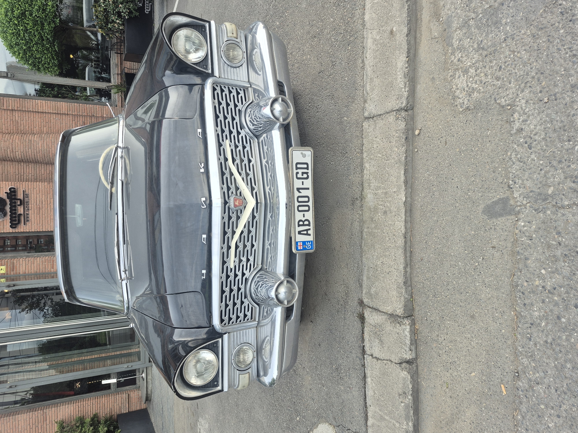 The image depicts the front of a black GAZ-13 Chaika vintage car parked near a building in Tbilisi, Georgia. The car has a distinctive chrome grille with the Cyrillic word "Чайка" (Chaika) spelled out across the top. A Georgian license plate "AB-001-GD" is prominently displayed on the front. The car is situated on an asphalt road adjacent to a concrete curb. The building behind the car appears to have large glass windows and a brick facade, possibly a restaurant or shop. There are some plants in front of the building. The overall setting is urban, suggesting a street in Tbilisi.