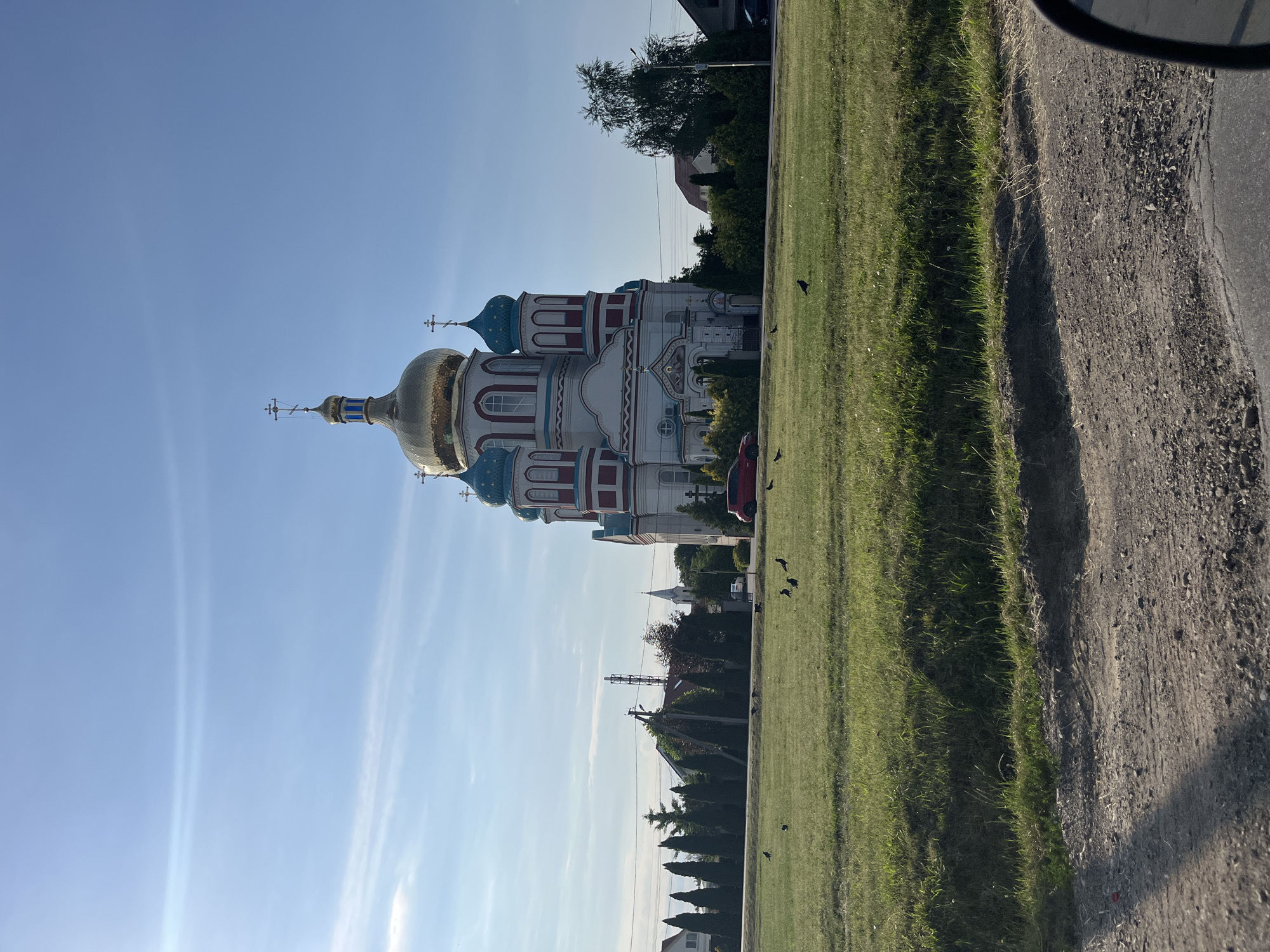 The image depicts a grand, multi-domed church in Mynai, Ukraine, bathed in the bright sunlight of a clear day. The church features a prominent golden dome and several smaller, turquoise domes, adorned with crosses. Its facade is white with red and blue accents, and it is surrounded by lush green grass, trees, and other buildings. A red car is parked in front of the church, and several black birds are scattered across the lawn. The foreground shows a rough, gravelly road surface, partially obscuring the view of the immediate surroundings. The sky is a pale blue with faint wispy clouds.