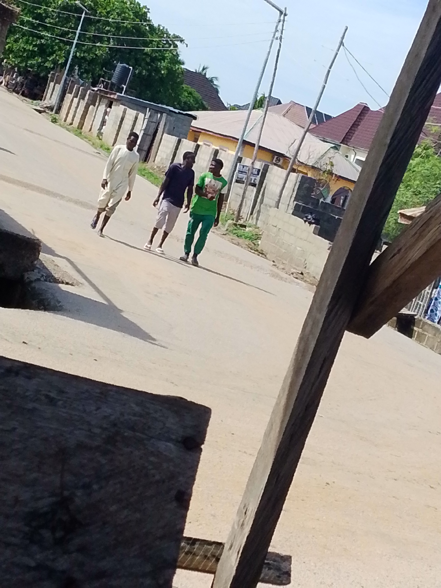 Three men are walking along a paved road in Lokoja, Nigeria. The man in the lead is wearing a light-colored robe, the second is in a dark shirt and lighter shorts, and the third is wearing a green shirt and pants. The road is bordered by block walls and buildings. Power lines run overhead. A wooden structure is partially in the foreground of the image, out of focus.