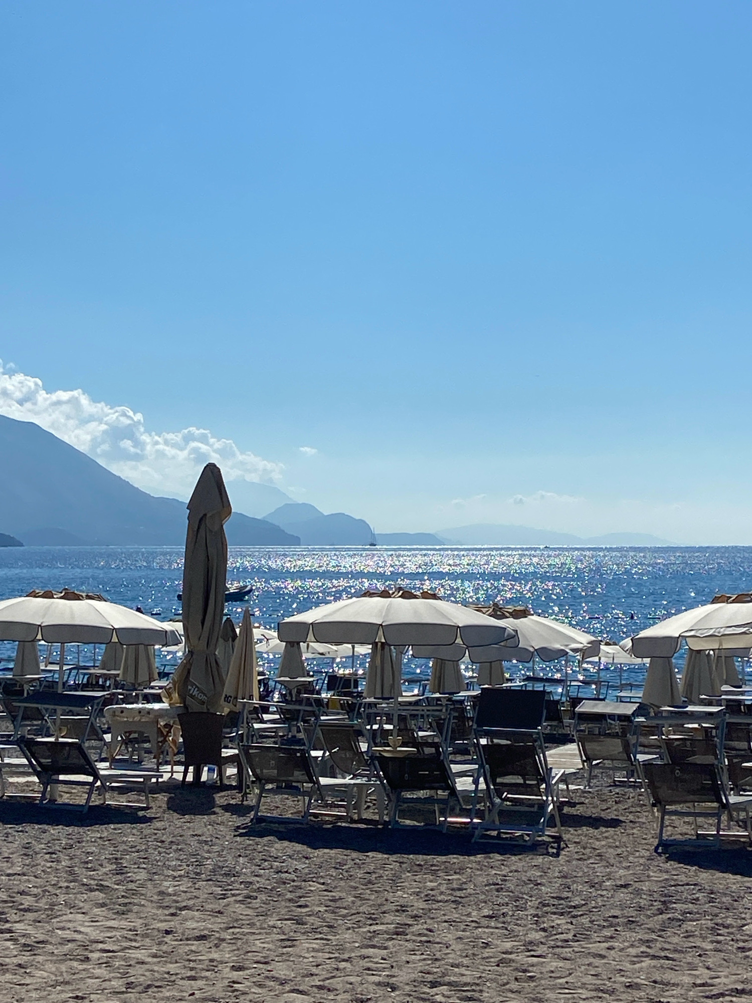 A serene beach scene unfolds under a clear blue sky, characteristic of Prijevor, Montenegro. In the foreground, a sandy beach is lined with rows of empty beach chairs and tables, partially shaded by closed white umbrellas. A tall, closed umbrella stands prominently in the center, with a small boat visible in the distance on the sparkling blue sea. Beyond the water, a chain of majestic mountains forms the horizon, their peaks softened by the atmospheric haze. The sea itself is a vibrant blue, reflecting the sunlight in a dazzling display of glitter. The overall atmosphere is tranquil, with no people present, suggesting an early morning or late afternoon setting.