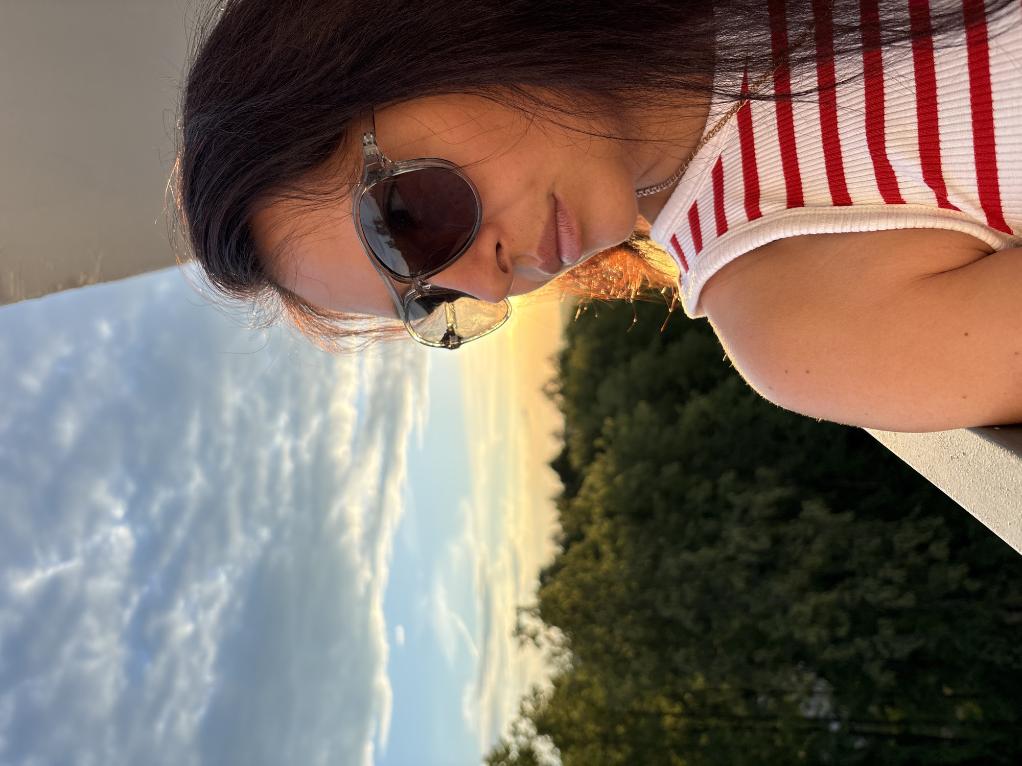 A young woman is looking down and to her right, with her head tilted. She has dark hair and is wearing sunglasses with clear frames and dark lenses. Her lips are slightly parted. She is wearing a white tank top with thin red stripes. A thin silver chain necklace is visible. The woman is outdoors, with a blurry background of trees on the right and a sky with clouds on the left. The sun appears to be setting or rising, casting a warm glow on the scene. The time of day is likely late afternoon or early evening. The lighting suggests a clear day. The provided location context is Chaikovsky, Russia. No visible text is present in the image.