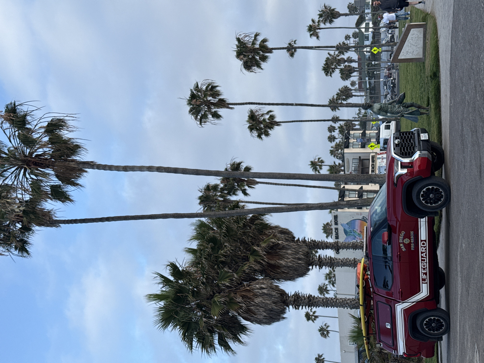 This image depicts a coastal scene in San Diego, United States of America, under an overcast sky. A prominent red lifeguard truck is parked on the right side of a paved area, facing the viewer. The truck clearly displays "SAN DIEGO FIRE-RESCUE" and "LIFEGUARD" in white text on its side. A yellow surfboard is secured on its roof rack.
Numerous tall palm trees, some with shaggy trunks and dense green fronds, line the left side of the scene and extend into the background. Behind these trees, various buildings are visible, including one with the text "BEACH HOTEL" and a colorful mural.
In the mid-ground, on a grassy strip, a dark green statue of a person, possibly holding a board or paddle, stands near a brown informational sign or monument. Several people are scattered on the paved path and grass, appearing to walk or stand casually. Yellow road signs, possibly indicating pedestrian crossings, are also visible further back.
The lighting is soft and diffused, characteristic of an overcast day. The scene portrays a typical, calm day in a San Diego beach community, with the lifeguard vehicle ready for duty.