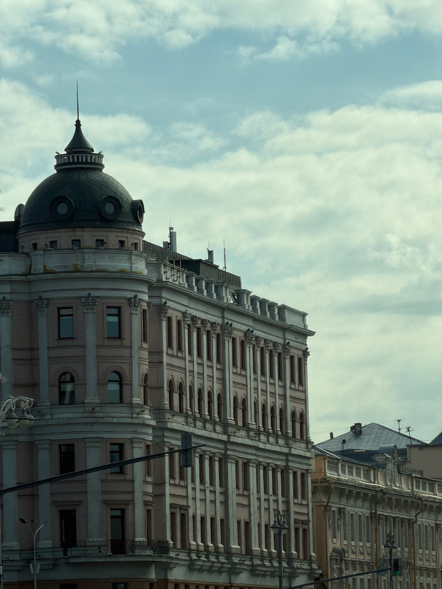 The image shows a view of a grand, light pink building with classical architectural details in Moscow, Russia, under a cloudy sky. The building features a prominent dark dome with a spire on its left side. The facade is adorned with rows of windows, columns, and decorative moldings. To the right of the main building, a portion of another building with similar classical elements is visible. The street level of the main building has arched windows and balconies. A street lamp with multiple lights is partially visible on the left side of the frame. A dark horizontal wire runs across the lower part of the image, possibly part of a traffic light or overhead power line. The scene captures a quiet urban streetscape, with no people or vehicles visible.