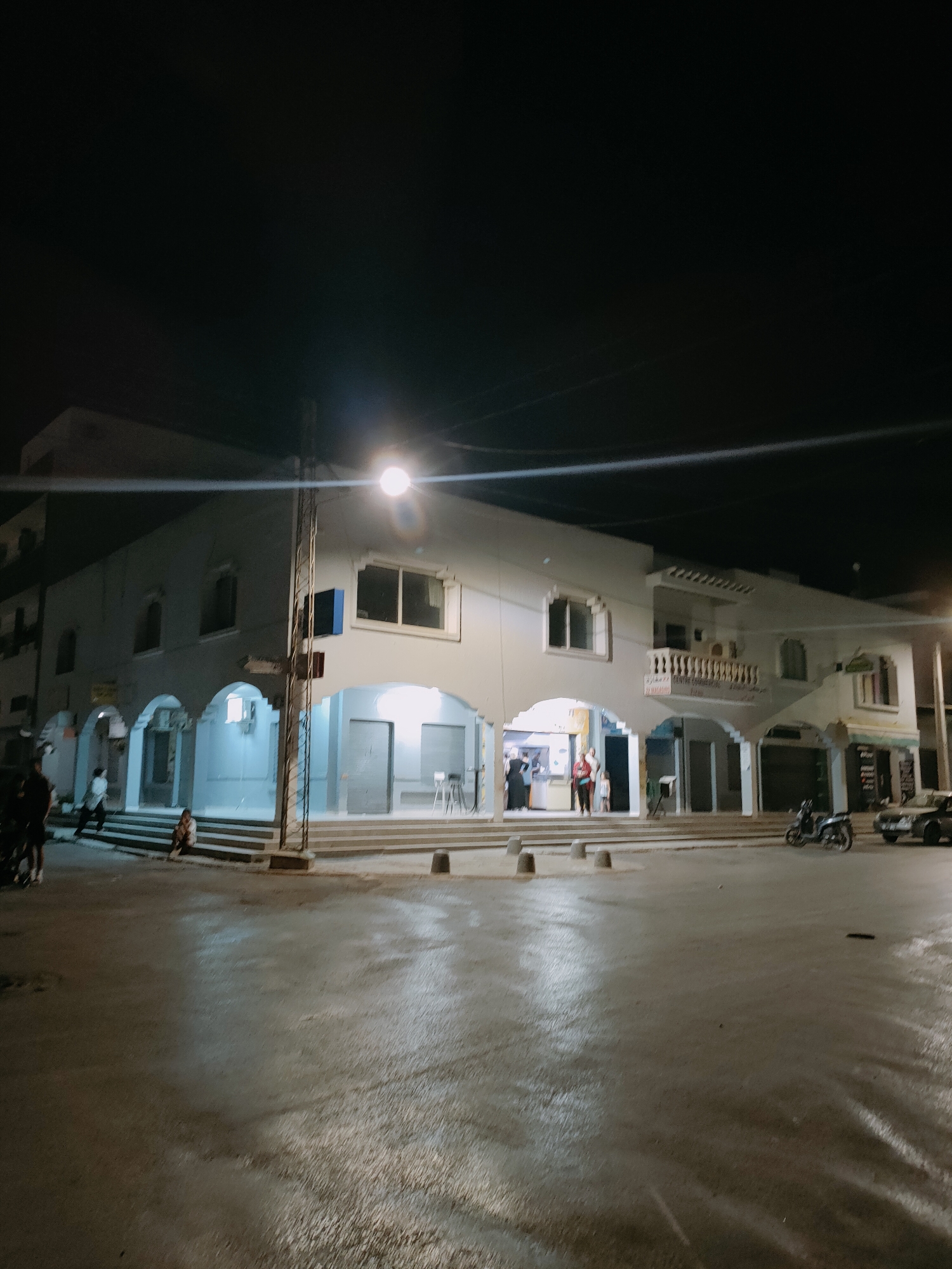 This image shows a two-story building at night, located on a street in Tunis, Tunisia. The building has a white facade with arched openings on the ground floor, which appear to be shop entrances or covered walkways. Several people are visible in front of the building, some standing and others sitting on steps. A motorcycle and a car are parked on the street in front of the building. The street appears to be wet, reflecting the ambient light from streetlights. The sky is dark, indicating it is nighttime. There are streetlights illuminating the scene. The architecture suggests a commercial or residential area. The overall atmosphere is that of a quiet urban street at night.