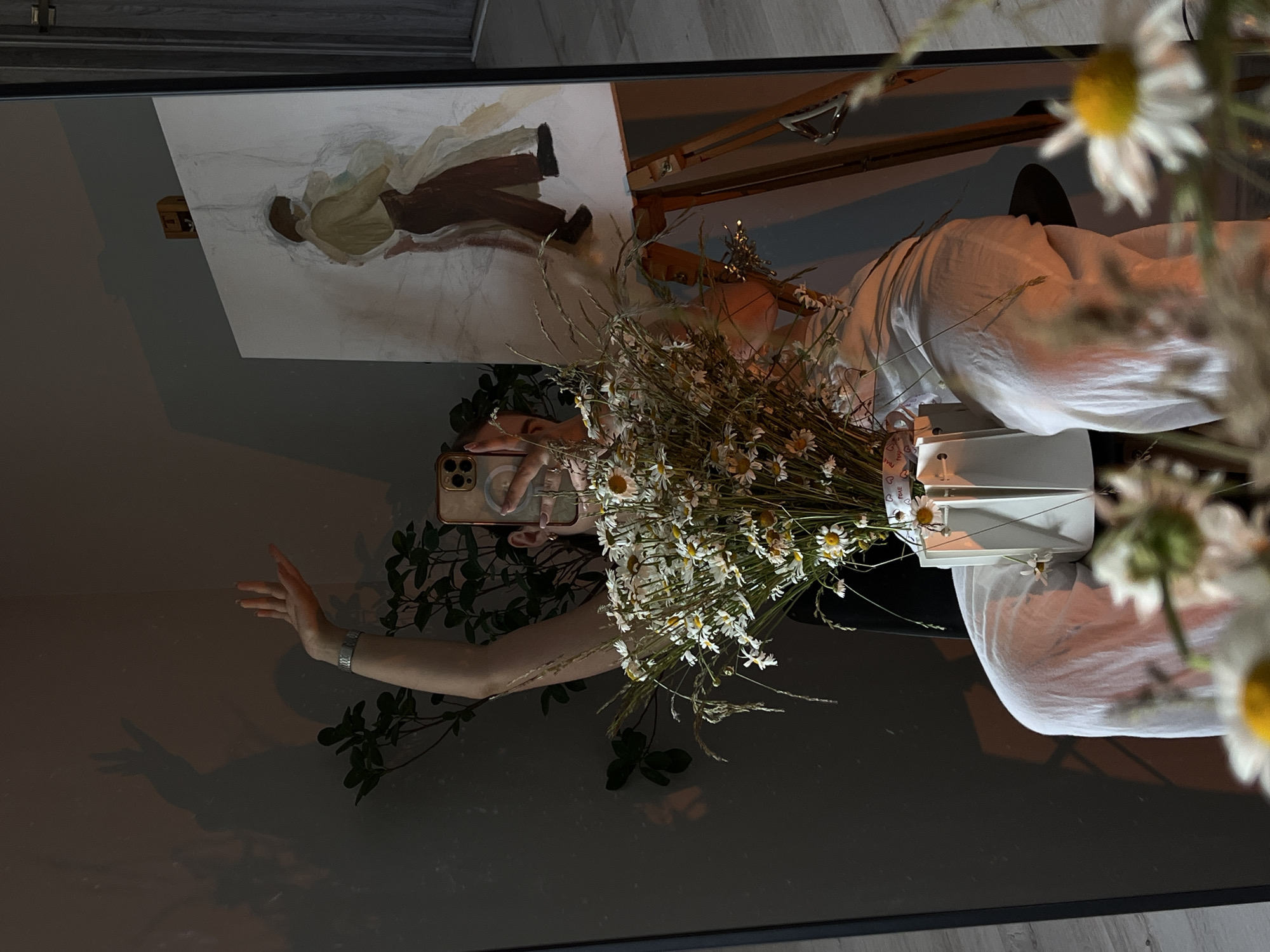 The image, taken indoors in Ternopil, Ukraine, shows a reflection in a mirror. A person is visible, holding a smartphone to take the photo, and a bouquet of daisies in a white vase. In the background of the reflection, an easel stands with a painting of two figures. The scene is dimly lit, with warm light casting shadows on the wall. The person is wearing white clothing.