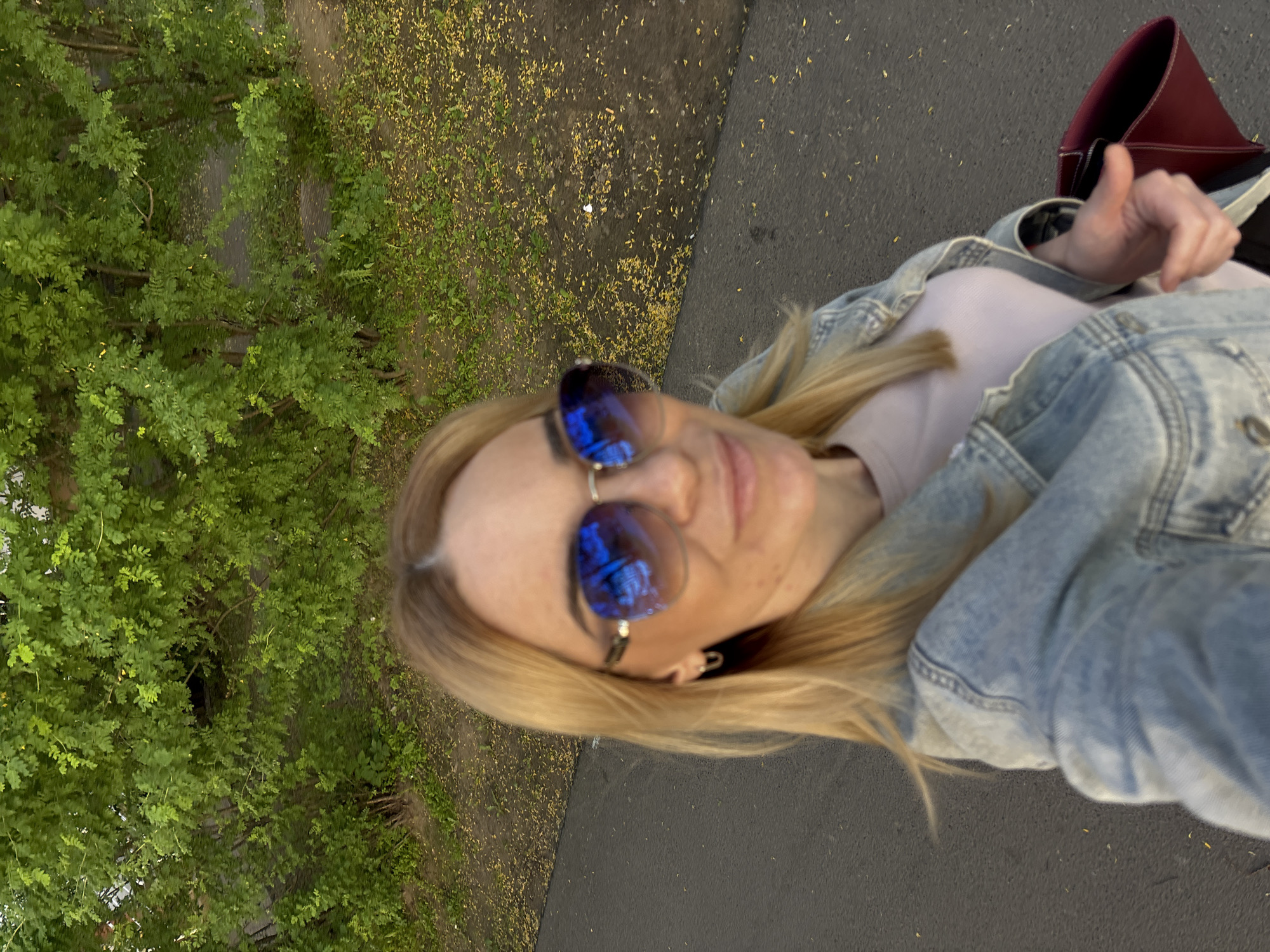 A blonde woman takes a selfie outdoors, giving a thumbs-up gesture. She is wearing a light pink top, a light blue denim jacket, and blue reflective sunglasses that obscure her eyes. Her hair is visible around her face and shoulder. A red bag is partially visible behind her right arm. The background features lush green foliage (likely trees or bushes) on the left side, and a dark paved surface (possibly asphalt) on the right. The ground in the middle, between the foliage and the paved surface, is covered with a scattering of small yellow debris, resembling fallen leaves or petals. The scene appears to be in natural daylight, suggesting it is daytime. No text is visible in the image.