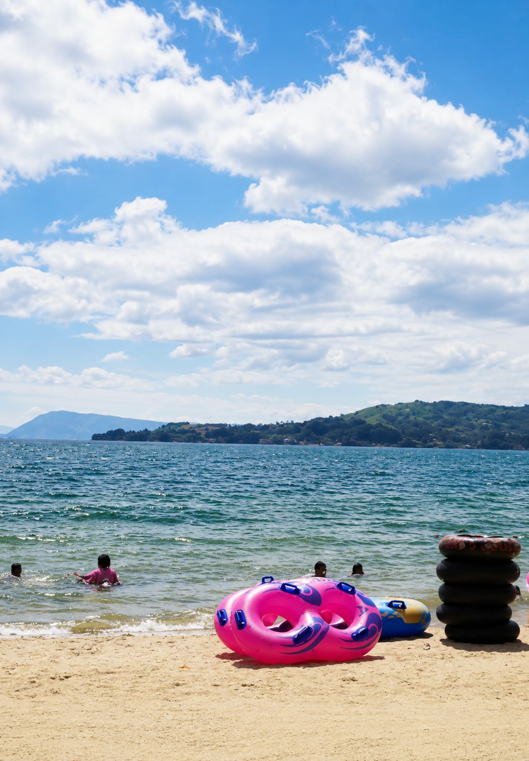 This image captures a serene beach scene at Lumban Bul Bul, Indonesia, under a bright blue sky dotted with fluffy white clouds. The foreground is dominated by a sandy beach with several inflatable tubes, one a vibrant pink with purple accents, and another a blue and yellow tube. To the right, a stack of black inner tubes stands tall. In the shallow, turquoise water, a few children are visible, some playing and wading, while others are partially submerged. The background features the expansive, shimmering water of a lake or sea, with a lush green, mountainous coastline stretching across the horizon. The overall atmosphere is one of peaceful recreation and natural beauty.