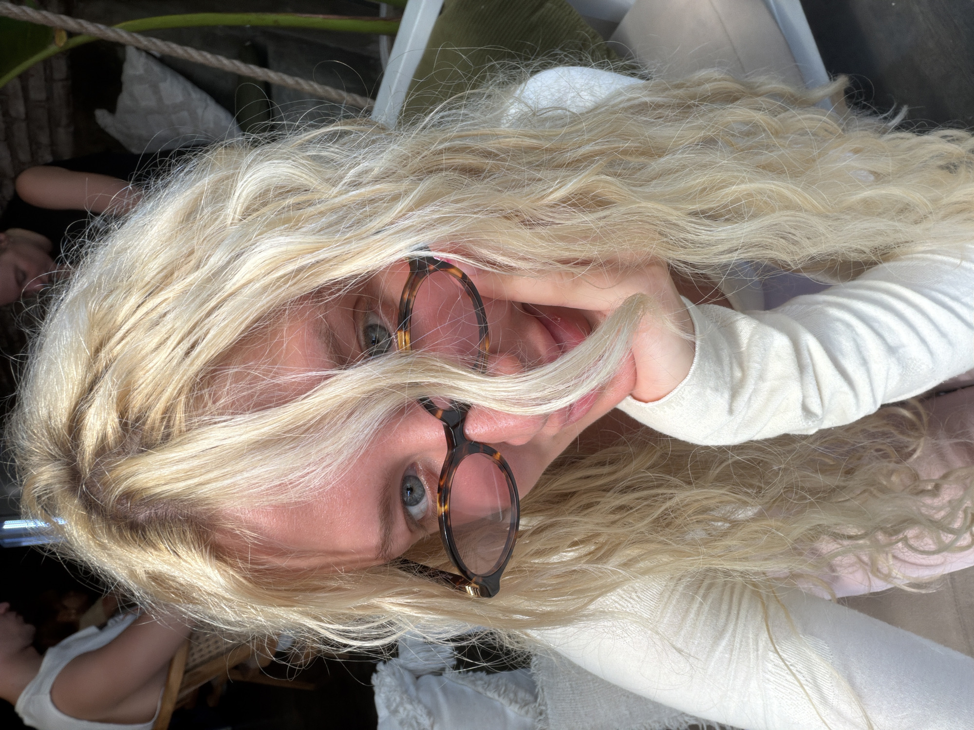 In a sunlit cafe in North Kuta, Indonesia, a young woman with long, wavy blonde hair and tortoiseshell glasses rests her chin on her hand. She is wearing a cream-colored sweater, and a pale pink garment is visible underneath. Sunlight streams through the scene, illuminating her face and hair. In the blurred background, two other individuals are seated at tables, one in a white sleeveless shirt and the other in a black sleeveless shirt. Cushions and greenery add to the relaxed, outdoor atmosphere of the cafe.