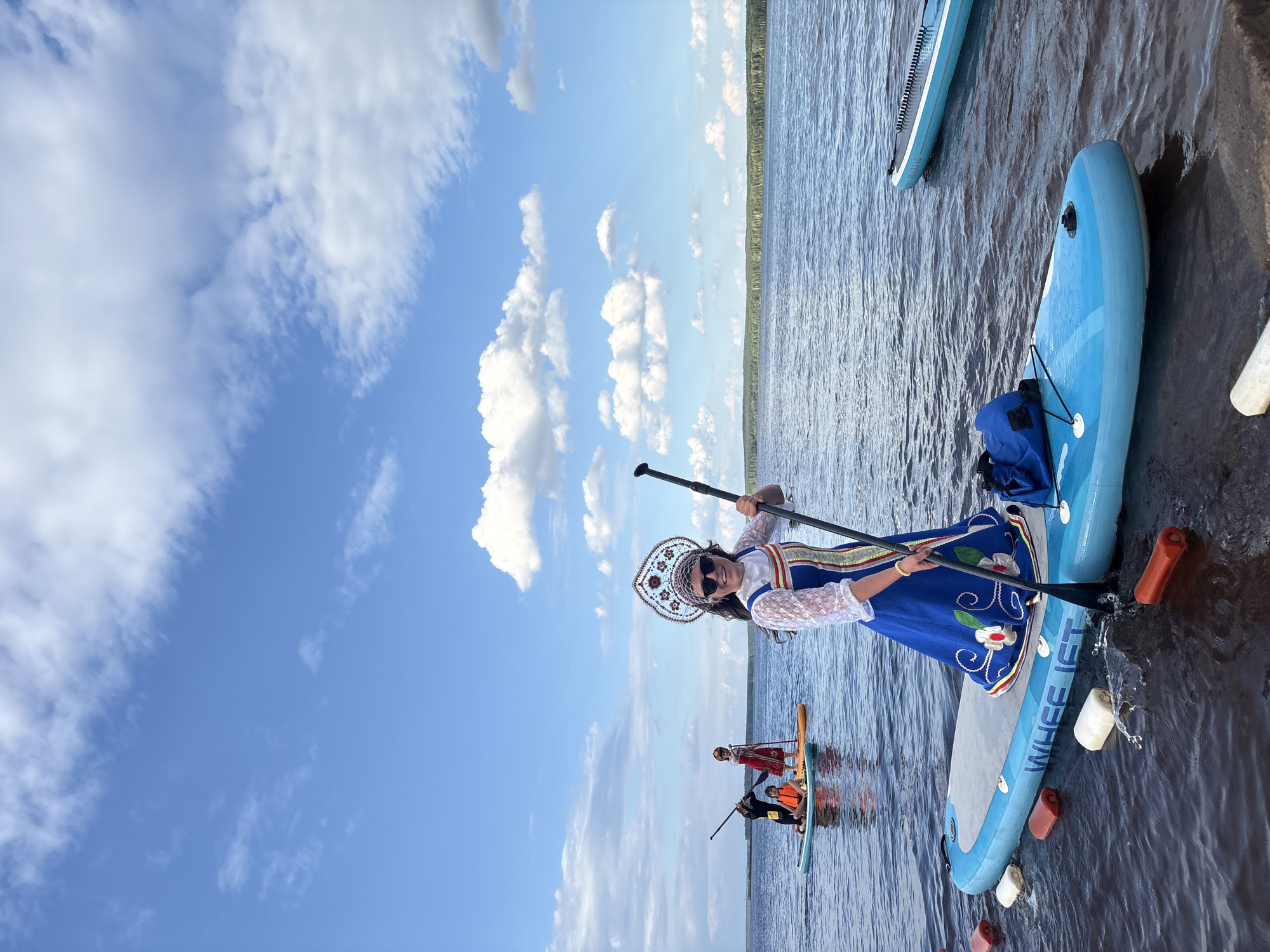 In Chaikovsky, Russia, a group of people is paddleboarding on a calm body of water. The main subject is a woman in the foreground, standing on a blue paddleboard. She is wearing a traditional Russian costume, complete with a decorative headdress and a blue dress adorned with floral embroidery. Her hair is dark, and she is wearing sunglasses. She holds a paddle in her hands, poised as if about to paddle or having just finished.
Behind her, two other paddleboards are visible with people on them. One paddleboard is orange and has a person wearing a red garment. The other paddleboard is turquoise with a person wearing a black top and an orange life vest. All individuals appear to be engaged in paddleboarding.
The scene is set under a bright blue sky with scattered white clouds, suggesting fair weather. The sun is casting light, creating reflections on the water. A treeline is visible in the distance along the far bank of the water, hinting at a natural or park-like setting. The water is calm, with ripples indicating gentle movement. The paddleboards are made of inflatable material, with one clearly displaying the brand name "WHEEL IS T" (likely a partial view of "WHEEL IS T...") on its side. The overall mood appears to be recreational and cheerful, with participants enjoying an outdoor activity on a sunny day.