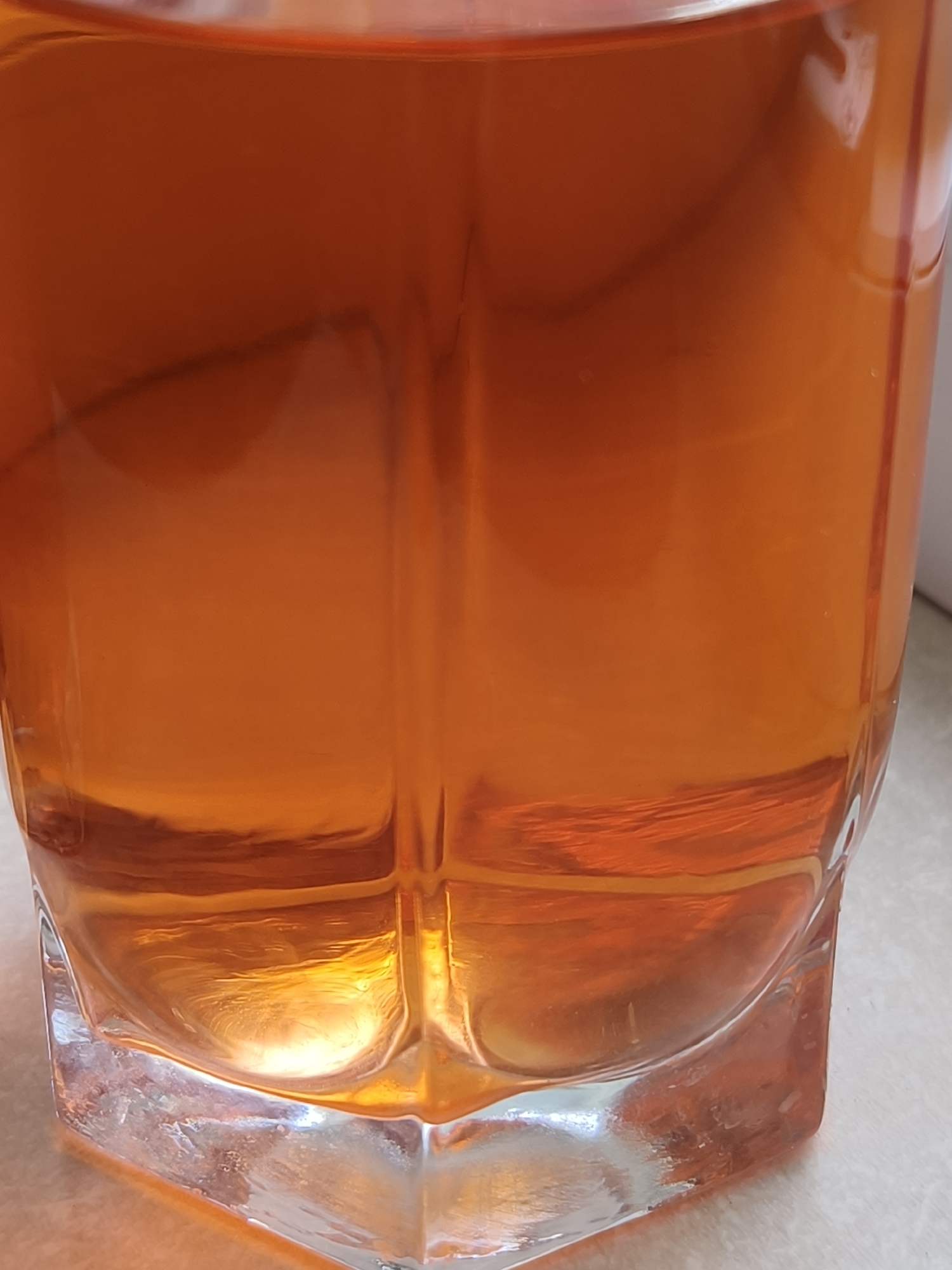 A close-up shot of a clear glass filled with an amber-colored liquid. The glass has a faceted base and straight sides with vertical indentations. The liquid appears to be still, with subtle internal refractions. The base of the glass shows some water spots or condensation. The surface underneath the glass is a light-colored, possibly tiled, surface. The context of Kryvyi Rih, Ukraine, is not apparent from this image.