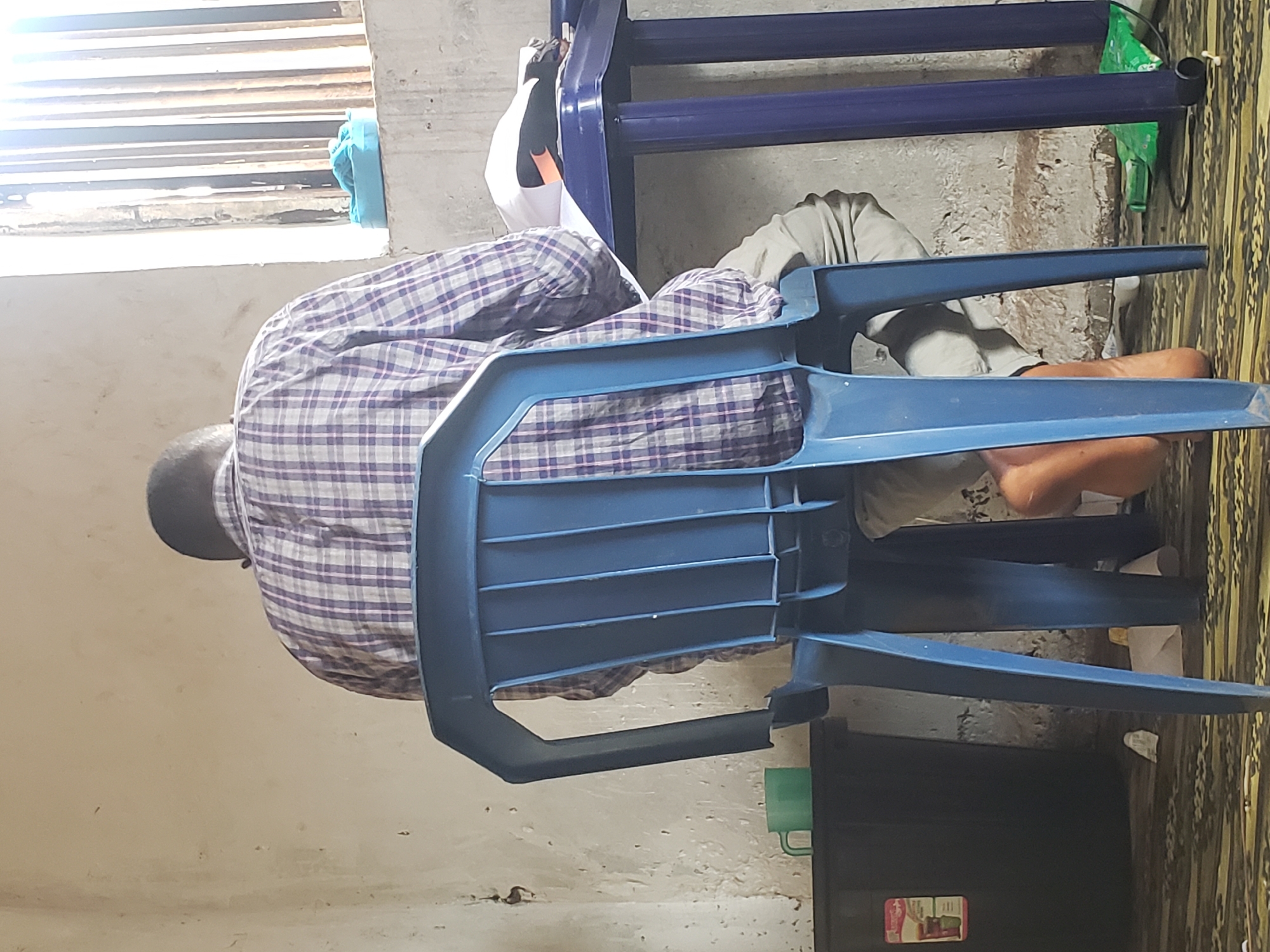 In Kachia, Nigeria, a person is seated at a blue plastic table, facing away from the camera, with their head bowed as if engaged in an activity like studying or writing. They are wearing a checkered long-sleeved shirt and light-colored trousers. The person is seated on a blue plastic chair. The table is covered with papers and what appears to be a white cloth. In the background, a window with vertical bars allows sunlight to stream in, illuminating the scene. To the left of the table, a black plastic container with a label is visible, and a green mug sits on top of it. The environment appears to be a simple room with a concrete floor and textured walls.