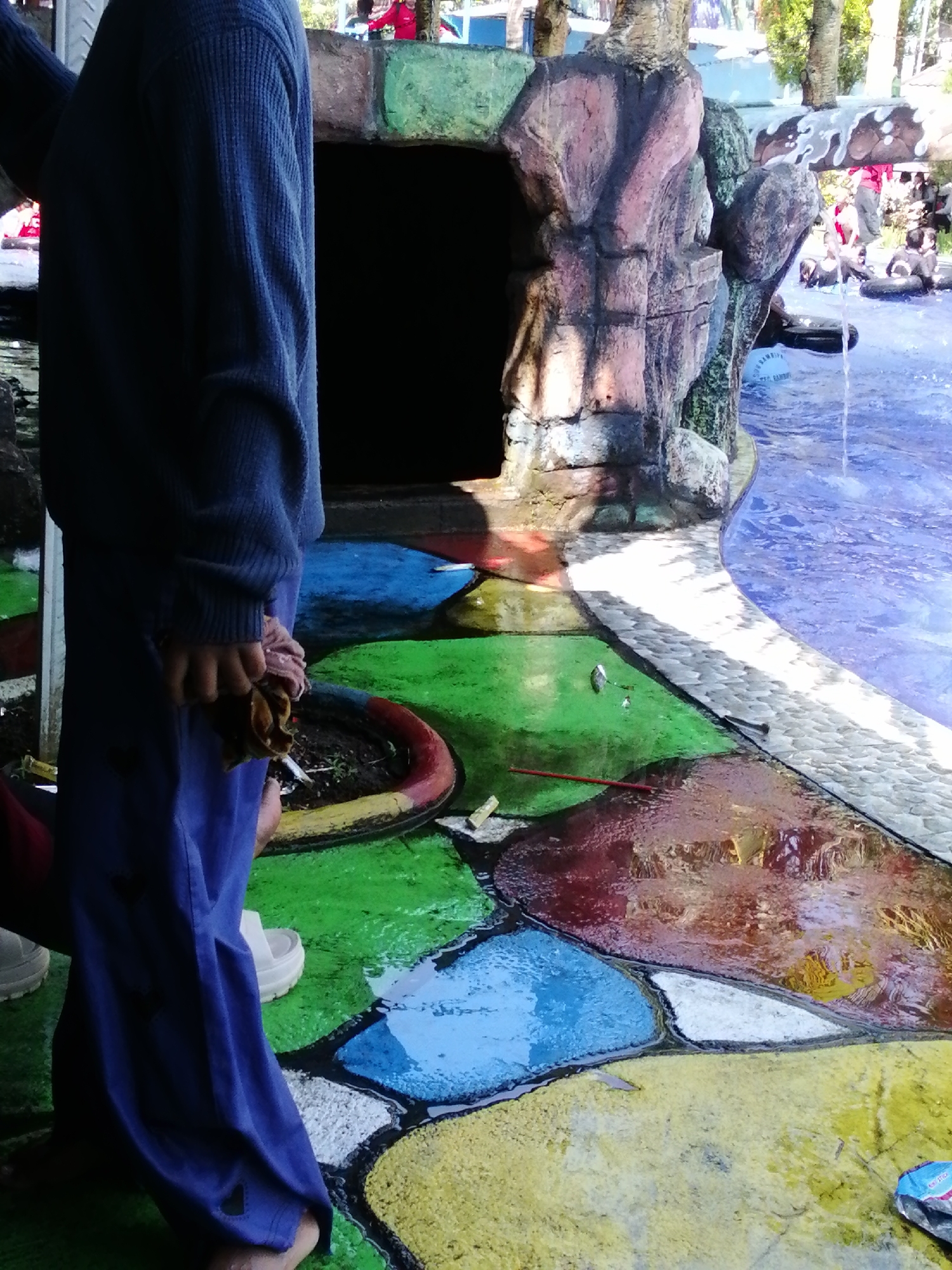 The image depicts an outdoor scene with a pool and a person standing in the foreground. The person is wearing a dark blue long-sleeved shirt and blue pants, holding a piece of cloth in their right hand. The ground around the pool is painted in a colorful, mosaic-like pattern of green, blue, yellow, and red. To the left of the person is a dark opening, possibly an entrance, with a rocky, artificial structure above it. In the background, to the right, is a pool of blue water where several people are visible floating on inner tubes. There are also trees and a structure with a white and brown striped roof in the distance. The overall impression is that of a public pool or water park.