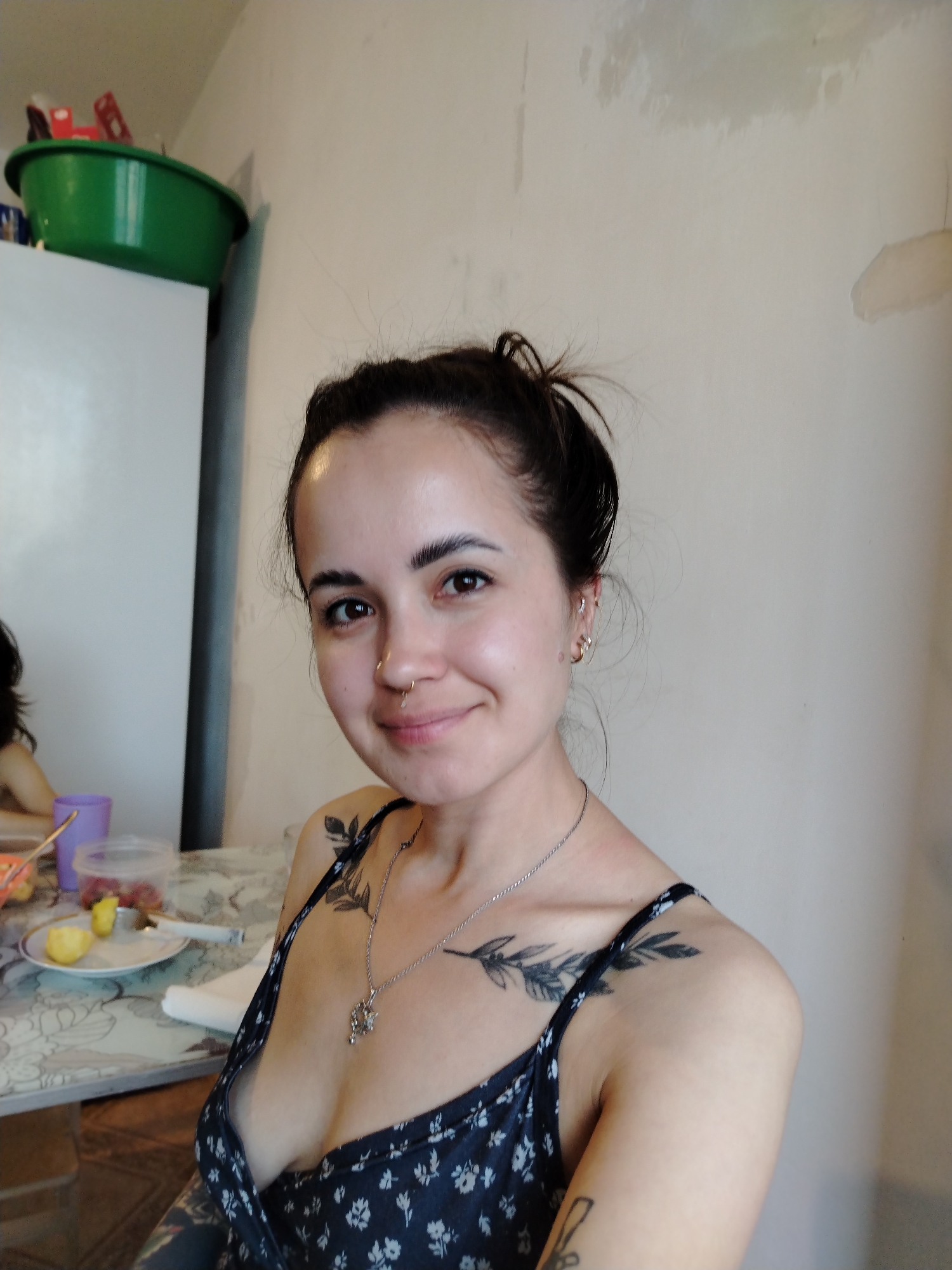 A young woman with dark hair pulled up in a bun and a nose ring smiles at the camera. She is wearing a dark blue and white floral print dress and a silver necklace. She has a tattoo of a leafy branch on her left shoulder and collarbone, and another tattoo on her arm.
She is sitting at a table with a white and grey patterned tablecloth. On the table are a plate with cut fruit, a purple cup, and a clear plastic container with some red fruit. In the background, to the left, is a white refrigerator with a large green basin on top, and some items inside the basin. The wall behind her is off-white and has some marks or stains on it. The scene appears to be indoors, possibly a kitchen or dining area. The lighting suggests it is daytime. The woman's expression is happy and relaxed. There is no visible text in the image.