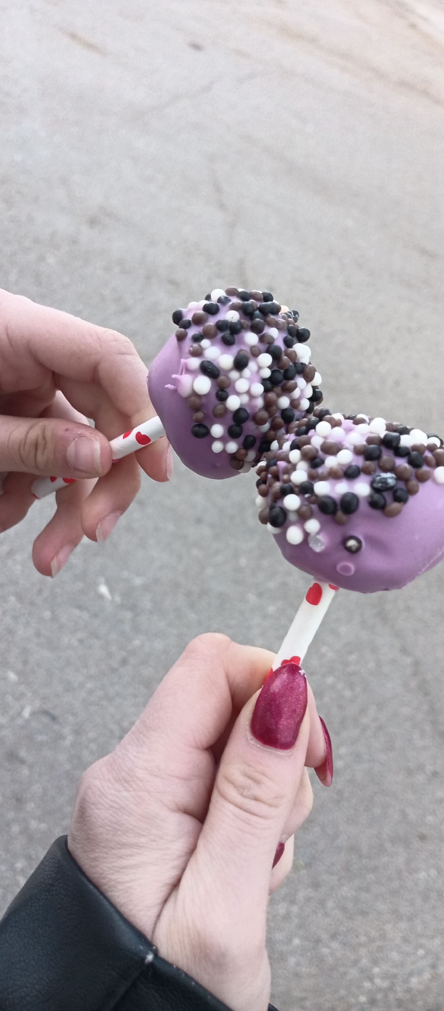 The image depicts two hands holding cake pops. The cake pops are lavender-colored and covered in white, black, and brown sprinkles. The sticks are white with red heart patterns. The hand on the right has long, burgundy-colored fingernails, and the person is wearing a black jacket. The background is a grey concrete surface. The image was taken outdoors. Location: Unknown, Unknown.