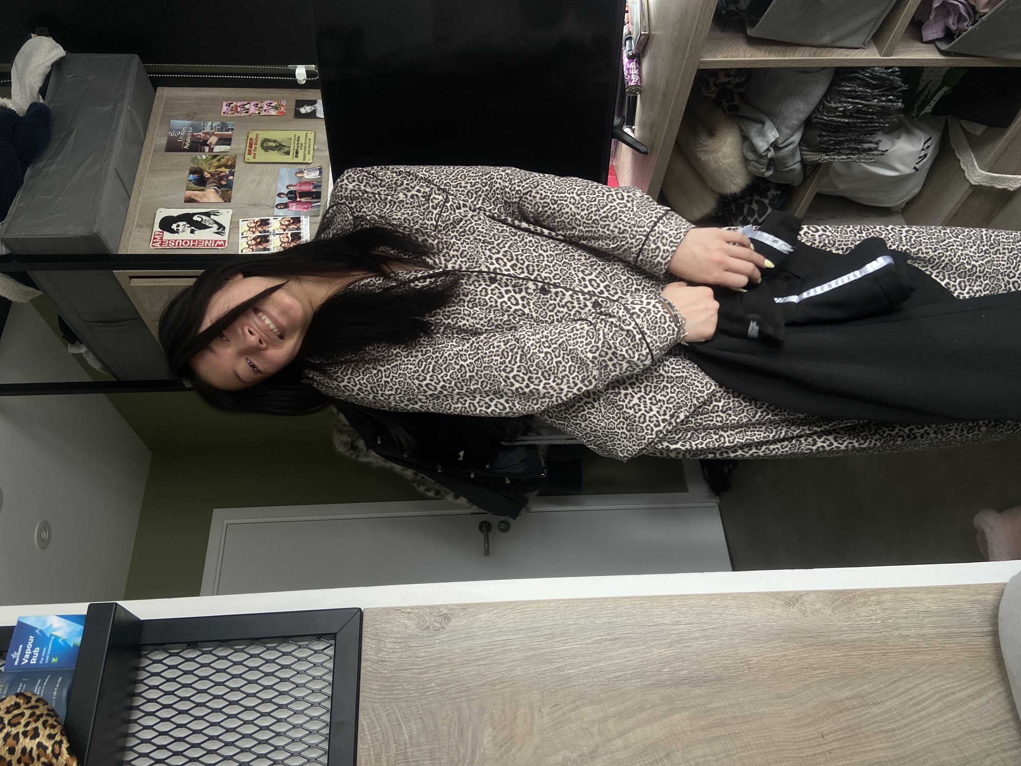 A smiling young woman with long, dark hair is shown in an interior setting. She's wearing a patterned pajama set and holding a pair of black pants with a white stripe. The background shows furniture and personal items, including storage shelves and a collection of images and memorabilia on a shelf behind her. The location is not discernible from the image.