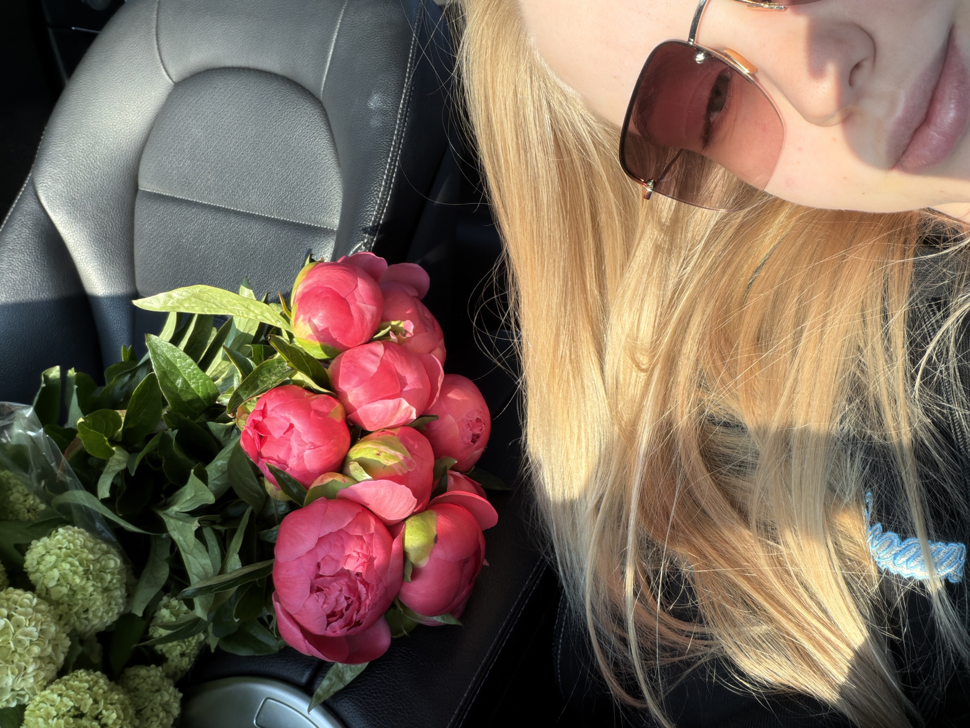 A close-up shot shows a woman with long, blonde hair and sunglasses, alongside a bouquet of pink and green flowers. The scene appears to be inside a car, as indicated by the car seat visible in the background. The location is undisclosed.