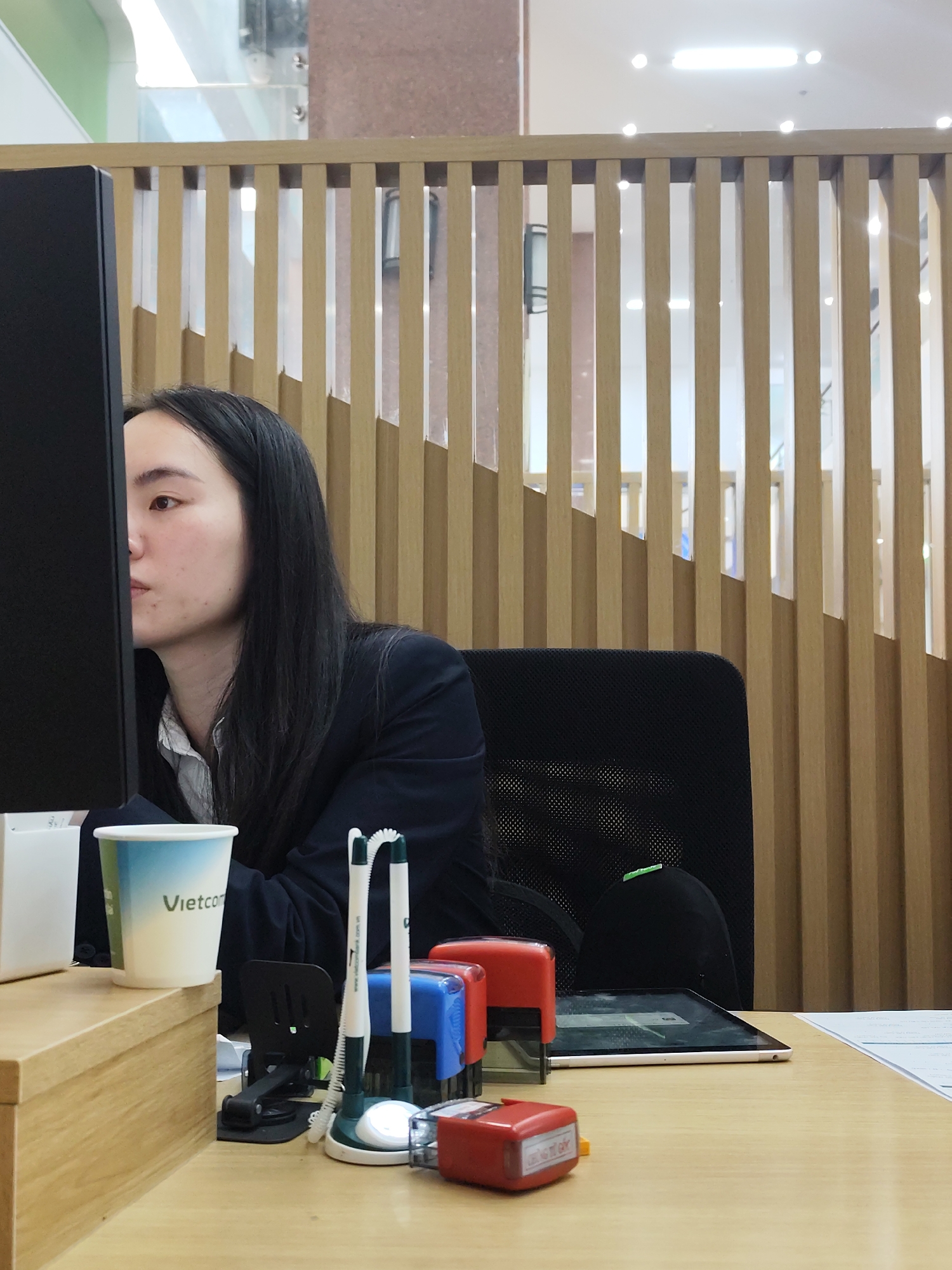 A young East Asian woman with long black hair, wearing a dark blazer over a white collared shirt, is seated at a wooden desk, looking intently at a computer monitor. The desk is cluttered with office supplies, including a white paper cup with "Vietcom" printed on it, two pens, a tablet, and two red self-inking stamps. A wooden partition with vertical slats forms the background. The scene suggests an office or administrative setting. While the image displays elements commonly found in Da Lat, Vietnam, such as the "Vietcom" branding (referring to Vietcombank, a major Vietnamese bank), there are no specific visual cues to confirm this exact location within the city.