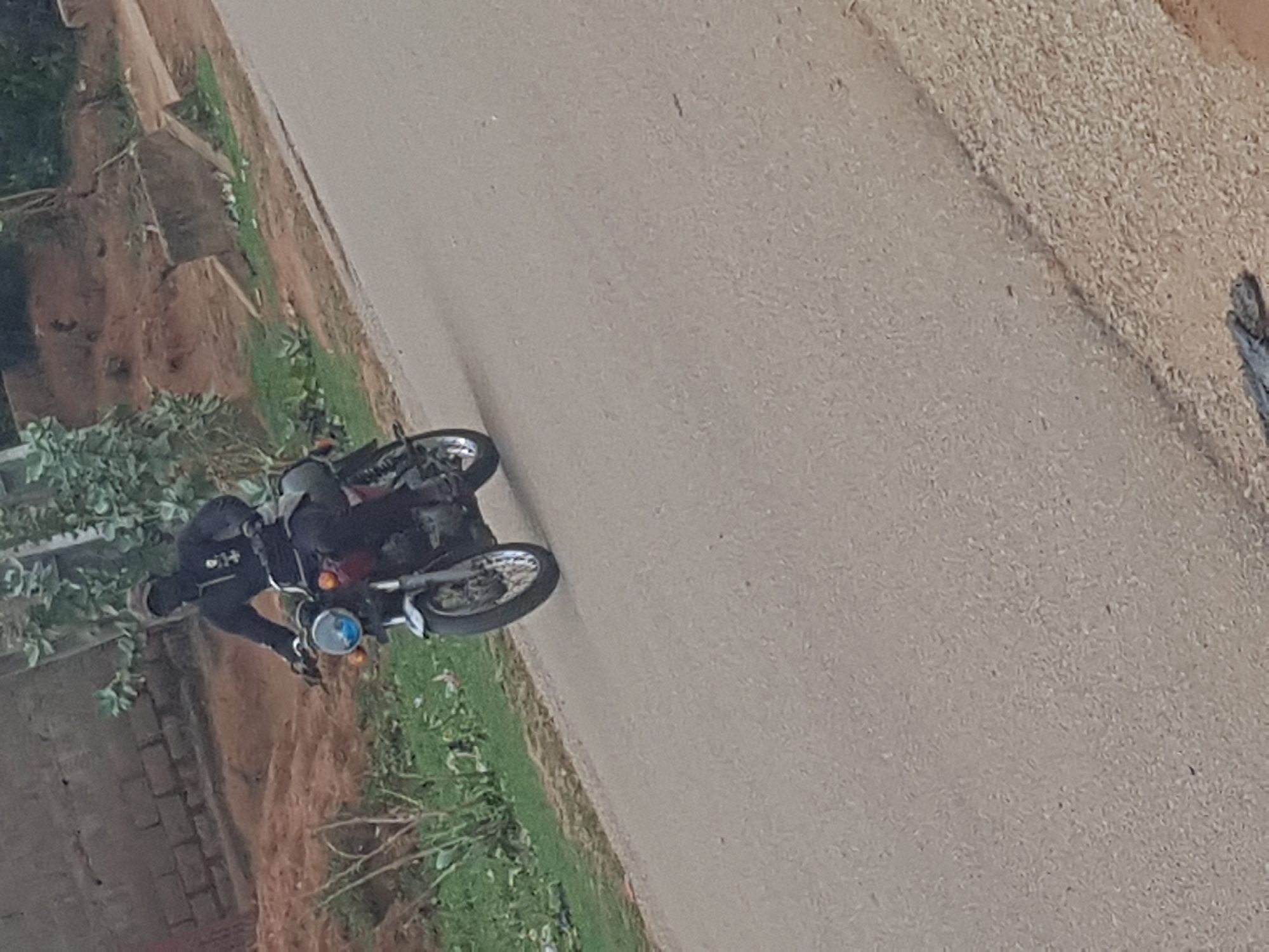 A motorcyclist is on a paved road in what appears to be Hanwa, Nigeria. The rider is wearing dark clothing and a helmet. The motorcycle is parked near the side of the road, with vegetation and a brick structure nearby. A dirt or gravel shoulder is visible to the right of the pavement.