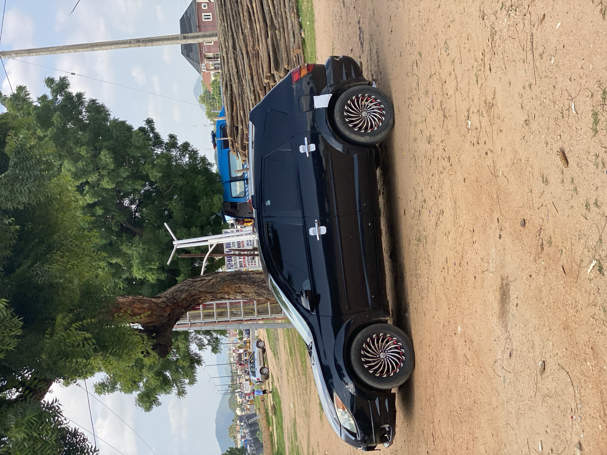The image shows a black car parked on a dirt road in Jalingo, Nigeria. The car has custom rims with a red and white pattern. In the background, there are buildings, a large tree, and a stack of logs. There are also other vehicles on the road, including a blue truck and a silver car. Signs are visible in the background with text that appears to be in English. The sky is cloudy, suggesting it might be daytime. The overall scene depicts a street or roadside environment in a town or city.