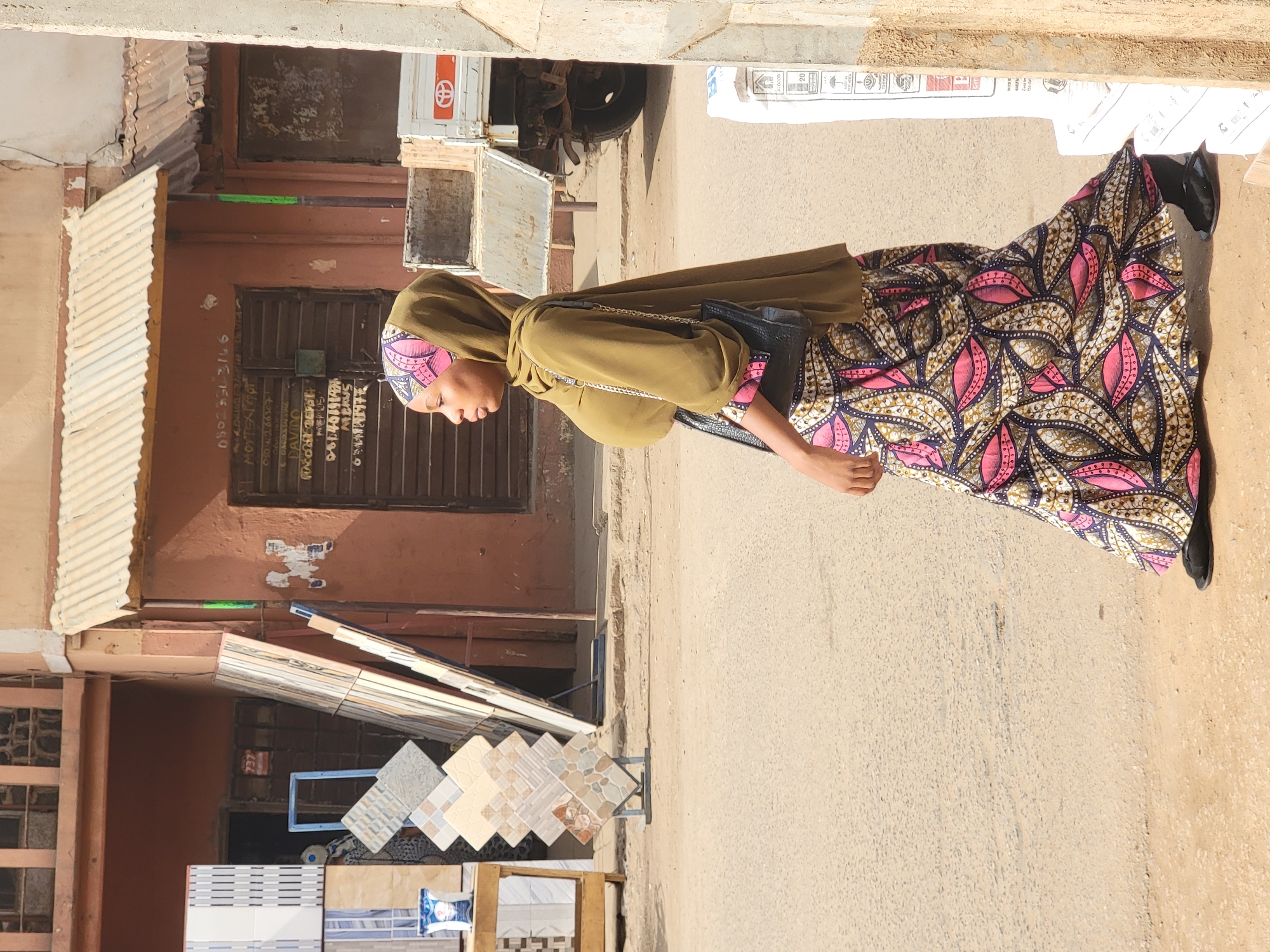 The image captures a daytime street scene in Maiburiji, Nigeria, under bright sunlight. The central subject is a woman standing or walking on a paved road, oriented to her left (viewer's right). She wears an olive green head covering (hijab or similar) and a matching long-sleeved top, paired with a striking, full-length skirt. The skirt features a vibrant pattern of pink, black, white, and gold/brown leaf-like or floral motifs. She carries a black shoulder bag with a chain strap and wears dark shoes or sandals.
To her left, a display of various floor tiles, including beige, grey, and patterned styles, is set up on metal stands, suggesting a shop selling building materials. Behind these displays are buildings with corrugated metal awnings and dark metal doors or shutters. On the facade of one brown building, several lines of handwritten text and numbers are visible, including "090-5643146," "MOHAMMED T," "080 42 6...", "DAVID," "MERYEM," and "MEHOS," likely indicating contact information or names.
In the background, on the opposite side of the street, a white truck or utility vehicle with a red circular logo (resembling Toyota) is visible. White sacks, some marked "20KG," are stacked against a wall, possibly containing cement or other construction materials. The clear, strong sunlight creates distinct shadows, highlighting the sunny weather. The street itself appears paved and somewhat dusty. The woman's gaze is directed downwards, and her expression is not clearly discernible.