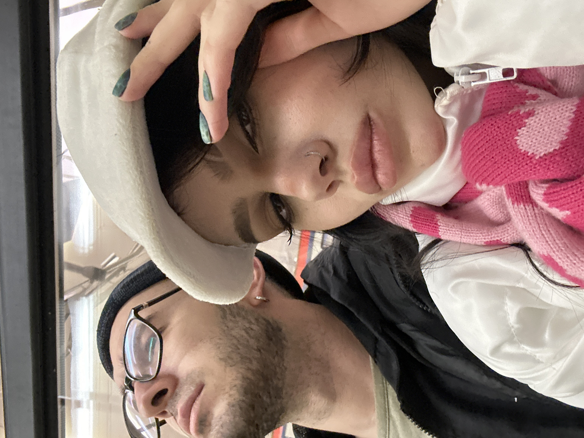 The image depicts a selfie of two individuals. A female is centrally positioned in the upper portion of the frame, wearing a white head covering and a pink and white knit scarf, with her hand touching her head. She has dark hair, a nose piercing, and her nails are painted a dark, iridescent green.
Below her is a male, partially visible, wearing a black beanie and glasses. He has light skin and a beard. The location appears to be indoors, with a window visible in the background. The overall orientation of the photograph suggests it was taken with a mobile device. Without further context, I cannot determine if the location is in Moscow, Russia.