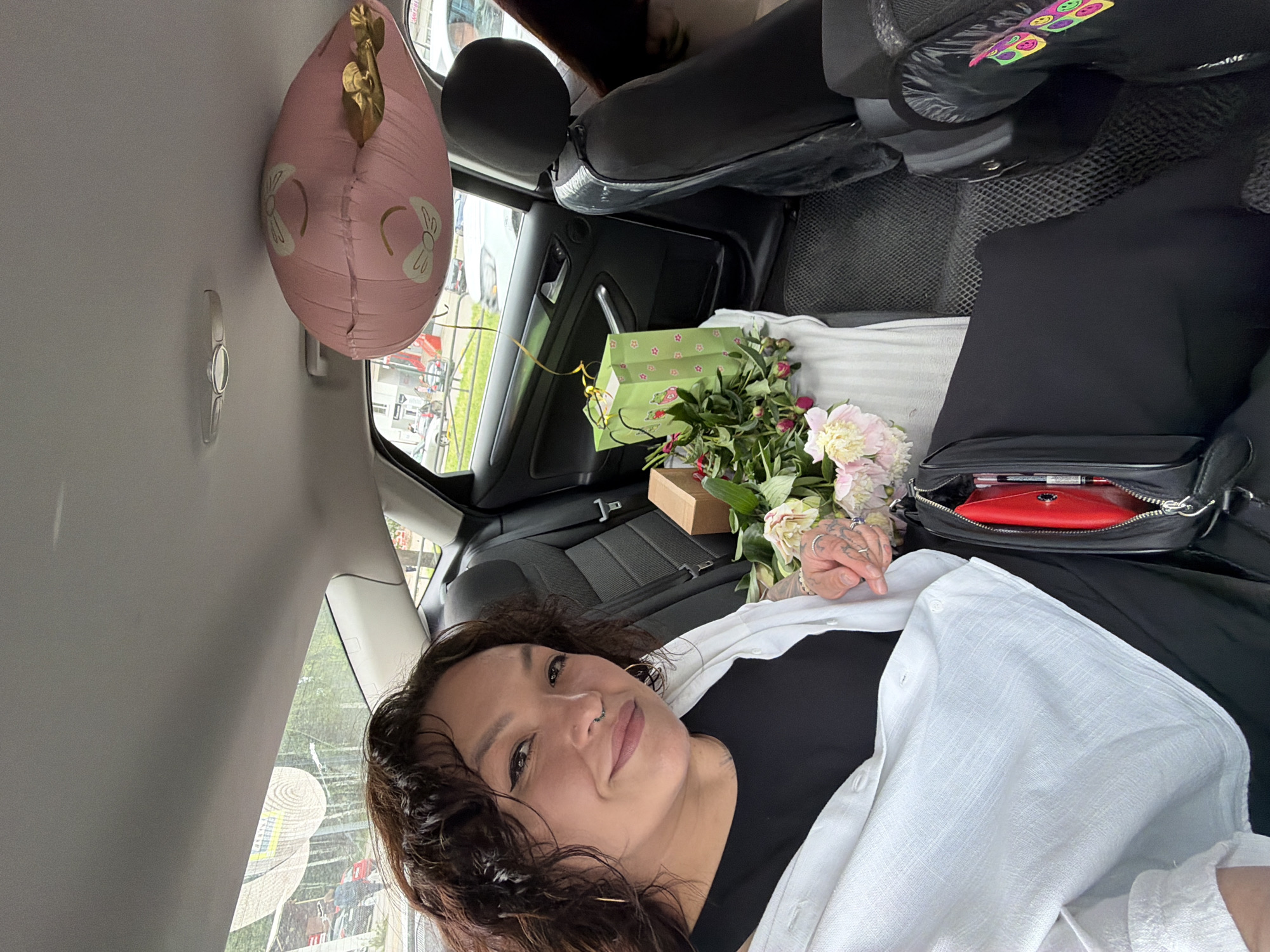A woman with curly dark hair, wearing a black top and a white open shirt, sits in the back seat of a car. She has a nose ring and multiple rings on her fingers. Beside her is a bouquet of pink and white peonies, a small brown gift box, and a green gift bag with strawberry designs. A large, pink, heart-shaped balloon with drawings of bows is attached to the car's ceiling. The car's interior shows black leather seats. Outside the car windows, a street scene is visible with buildings and moving vehicles. The image is taken in Chaikovsky, Russia, as indicated by the text on the yellow street sign visible in the background.