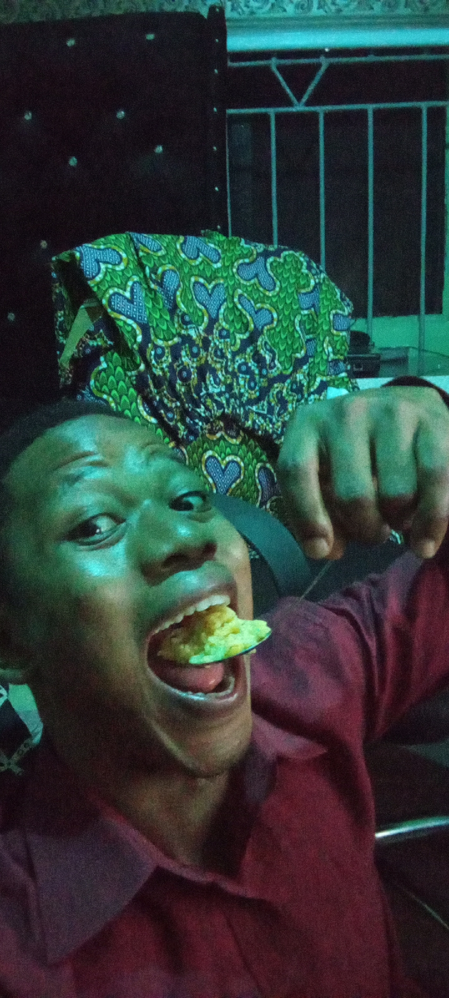 A close-up shot depicts a man with dark hair and skin, wearing a maroon-colored button-down shirt, holding a spoonful of yellow food near his open mouth. He is looking directly at the camera. In the background, there is a dark headboard with light-colored embellishments, a fabric with a green and blue heart pattern, and a metal railing. The location appears to be indoors in the city of Unknown, Unknown.