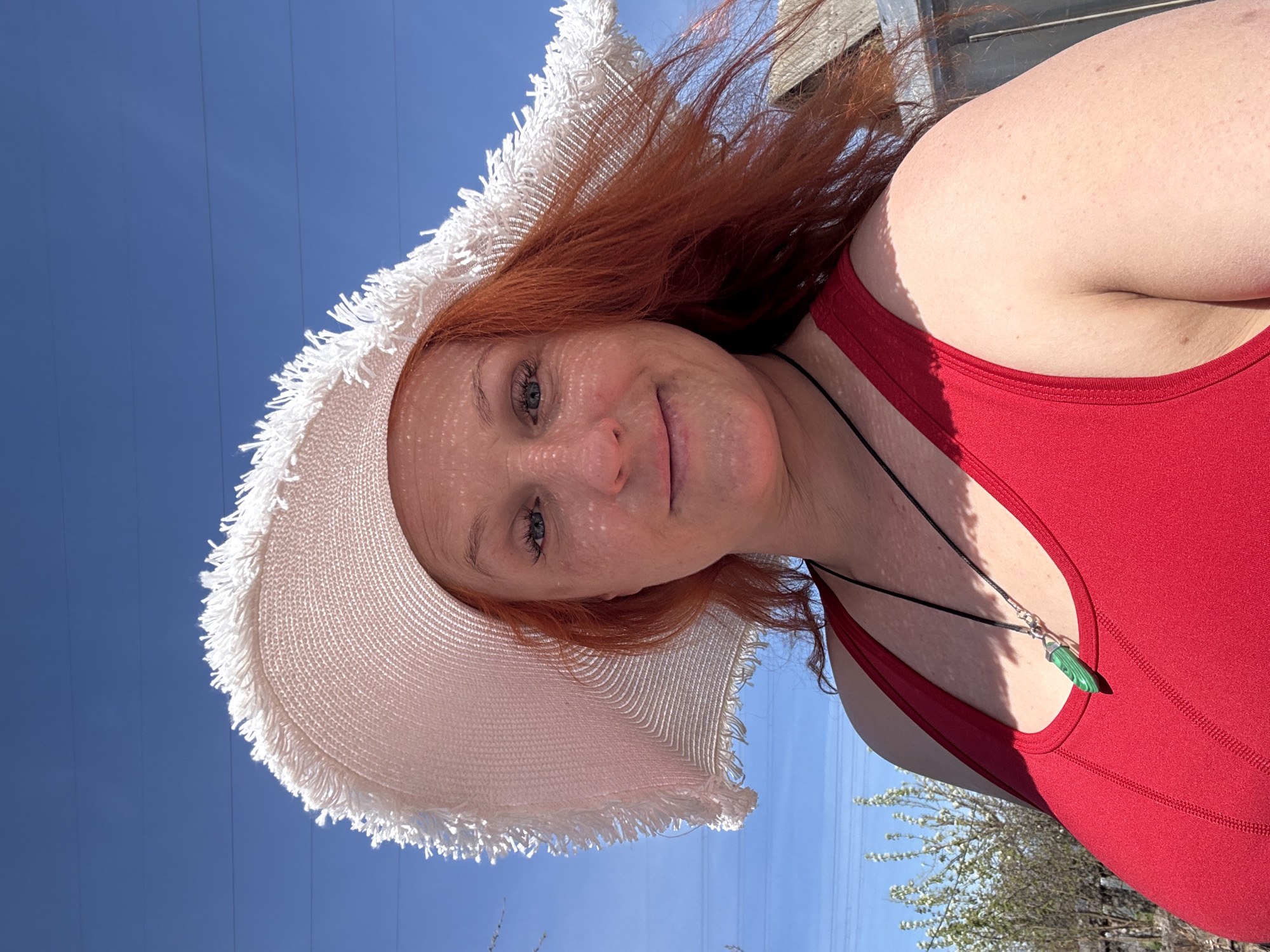 A woman with red hair wearing a white sun hat and a red tank top looks directly at the camera. She is wearing a necklace with a green pendant. The background includes a clear blue sky and some trees or bushes. There are also some electrical wires visible in the upper left corner. The location in the city of Chaikovsky, Russia is not discernible from the image.