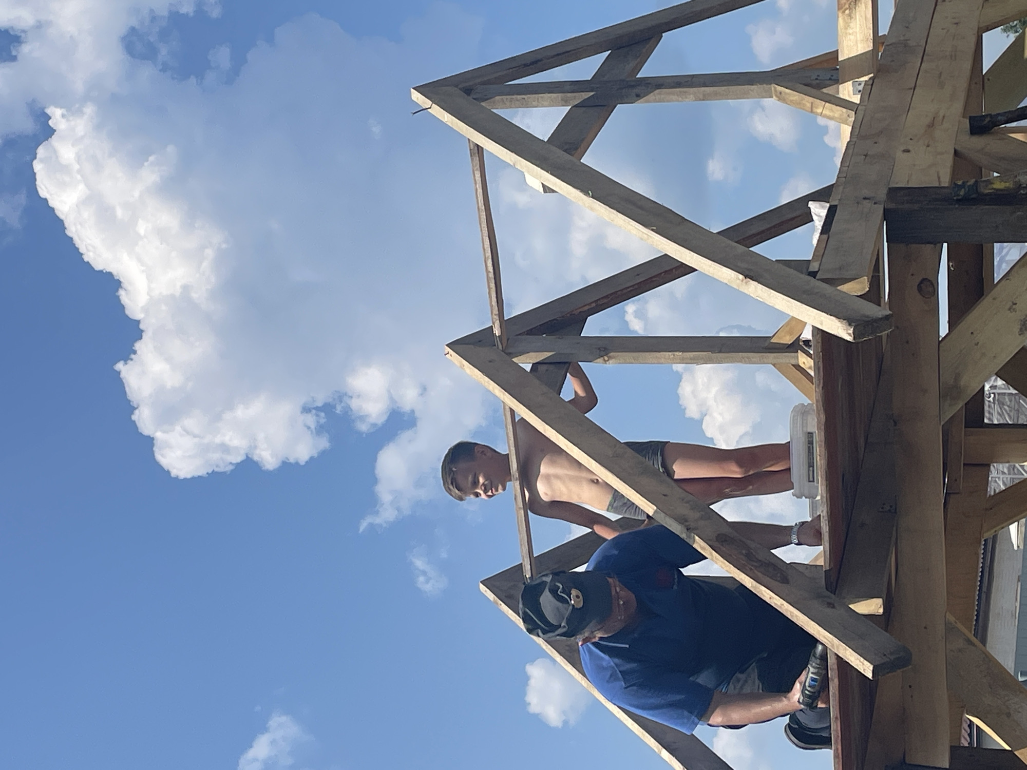 In the city of Maminskoe, Russia, two individuals are engaged in construction work on a wooden structure under a bright blue sky with scattered white clouds. A man in a blue shirt and dark pants, wearing a black cap with a circular emblem, is on the lower level of the structure, holding a cordless drill and appearing to be fastening a wooden beam. Beside him, a young boy, shirtless and wearing grey shorts, stands on a higher level of the structure, looking to his right with a smile. The structure is partially built, with exposed wooden beams forming the framework of what appears to be a roof. The overall scene suggests a cooperative effort in outdoor construction.
