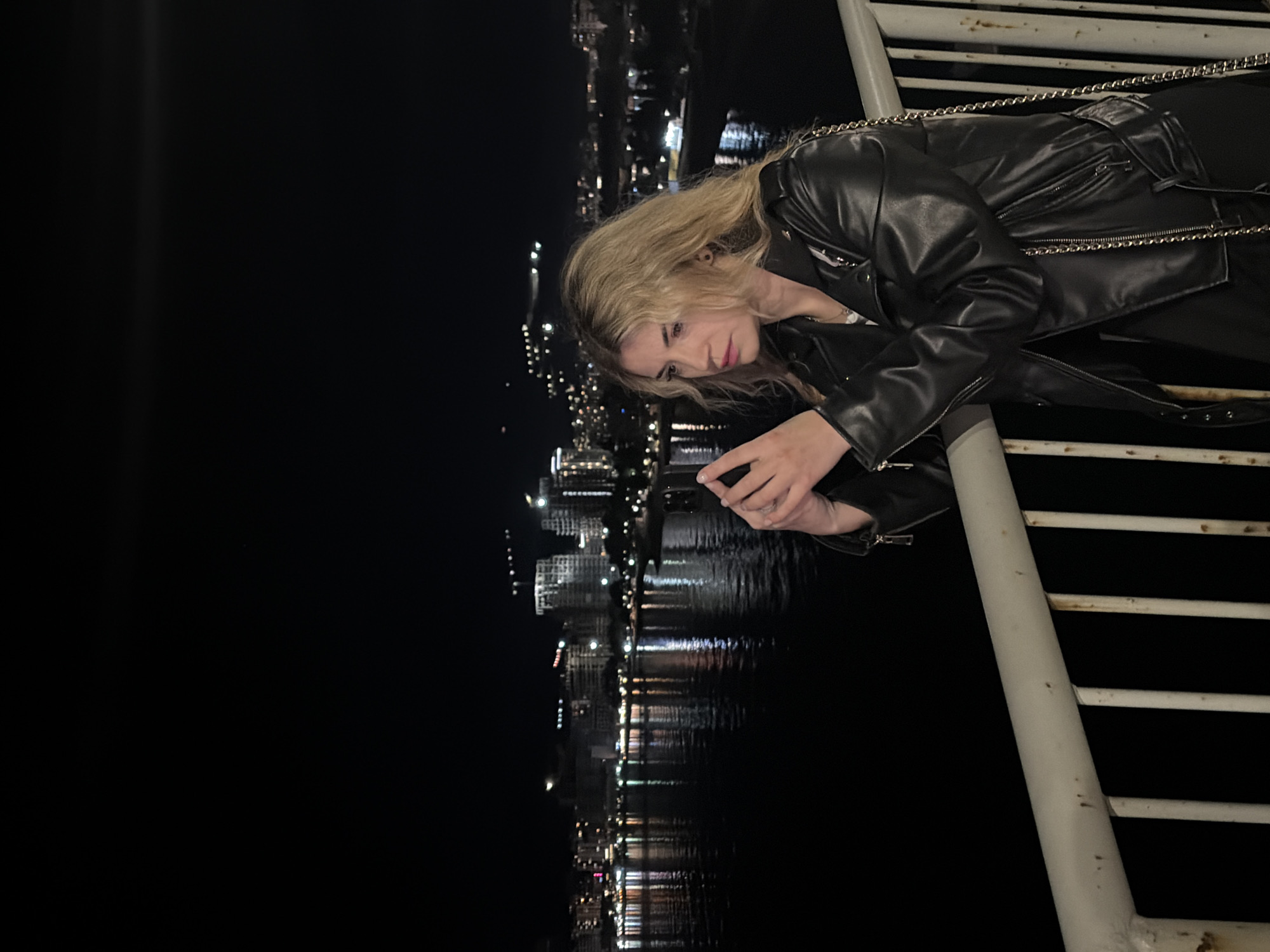 A woman is leaning over a white painted railing at night. She has long blonde hair and is wearing a black leather jacket. She is holding a cell phone in her hands and looking at it. The city of Budva, Montenegro, is visible in the background, with the lights reflecting on the water.