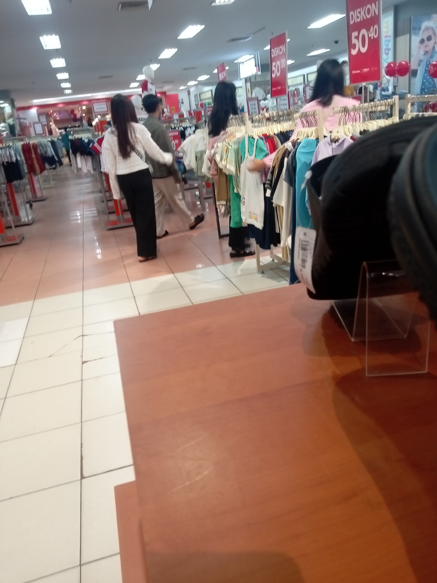 A wide-angle interior shot captures a retail store, likely a department store or supermarket, in Batam, Indonesia, evident from the "DISKON" signs indicating discounts in Indonesian. The lighting is bright and overhead, typical of commercial spaces. In the foreground, a polished wooden surface occupies the lower right, obscuring a portion of the scene. Behind this, a dark object rests on a transparent stand, also partially obscuring the view. The middle ground reveals aisles of clothing racks, with garments hanging on display. Several individuals are present. Towards the left, two women are walking away from the camera, one in a white sweater and black pants, the other with dark hair. Further back, a man in a dark jacket and a woman with dark hair are also visible. In the background, more clothing racks and promotional signs are evident, including red banners with "DISKON 50% 40" and a "mizzy" logo. The floor is tiled, with a mix of light and darker pink squares, showing some wear and tear. The overall impression is a busy shopping environment during a sale.