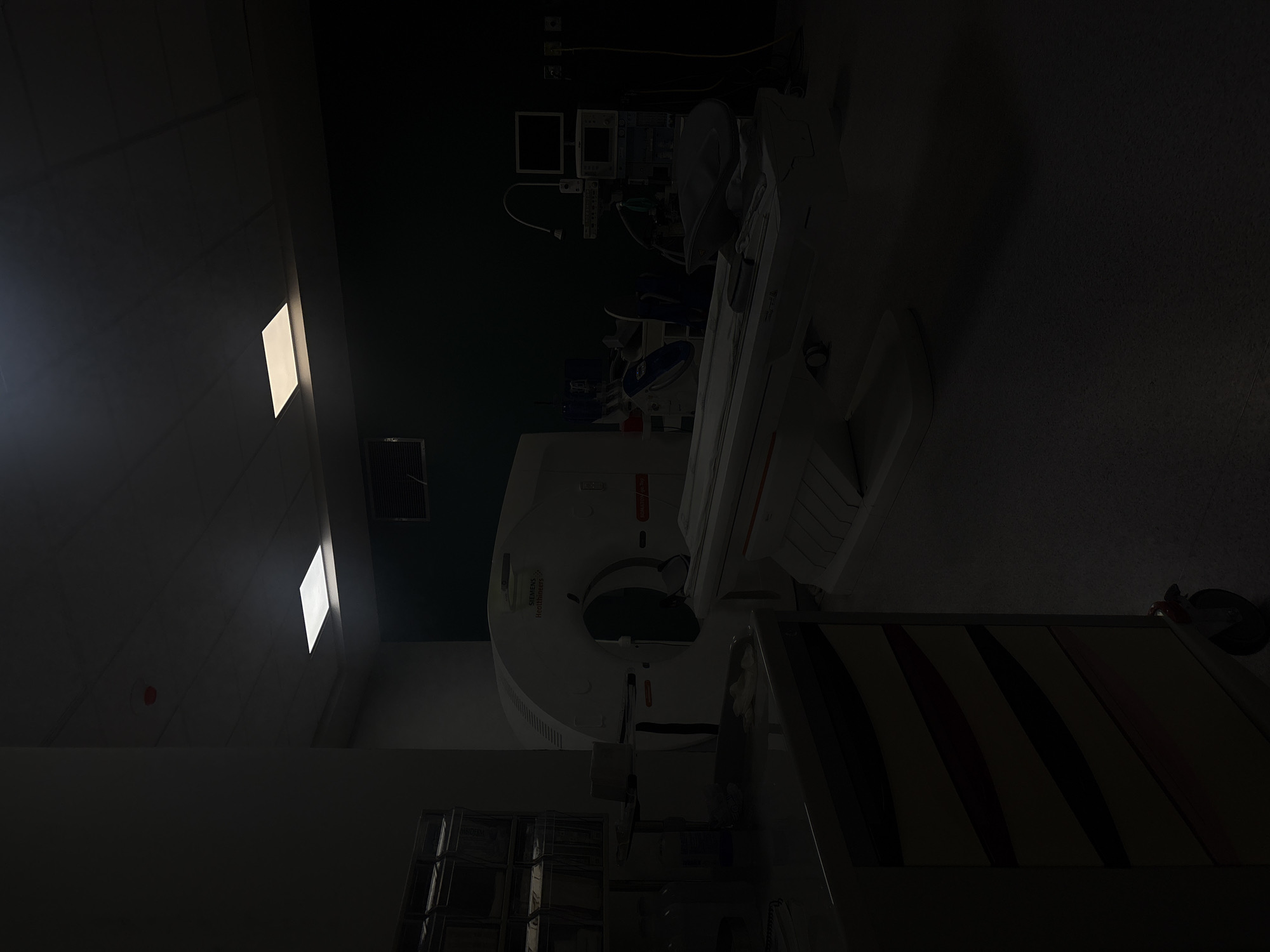 The scene depicts a dimly lit room, likely a medical facility, with a CT scanner as the central element. Various medical equipment is present, including monitors and other support devices. There is a medical cart in the foreground, partially obscuring the view. The overall impression is of a clinical environment prepared for medical imaging procedures. The location cannot be determined from the image alone.