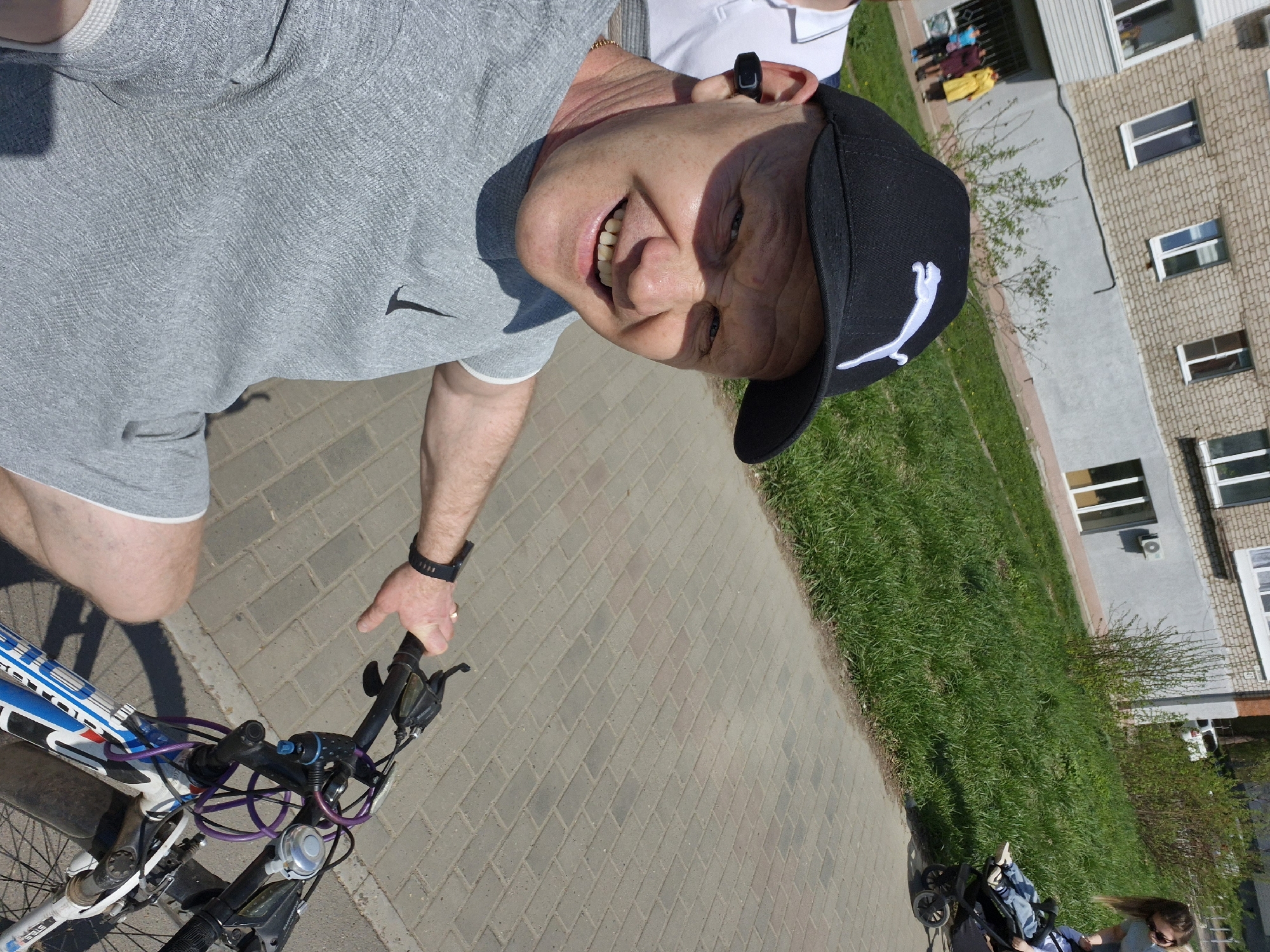 A man wearing a gray t-shirt, black shorts, and a black Puma baseball cap is riding a bicycle on a brick sidewalk. He is smiling at the camera. He also appears to be wearing a black smartwatch and a black electronic device in his right ear. The sidewalk runs alongside a grassy area. Beyond the grass is a multi-story building with numerous windows and air conditioning units. In the background, there are people and what appears to be a baby stroller. This image appears to be taken in Chaikovsky, Russia.