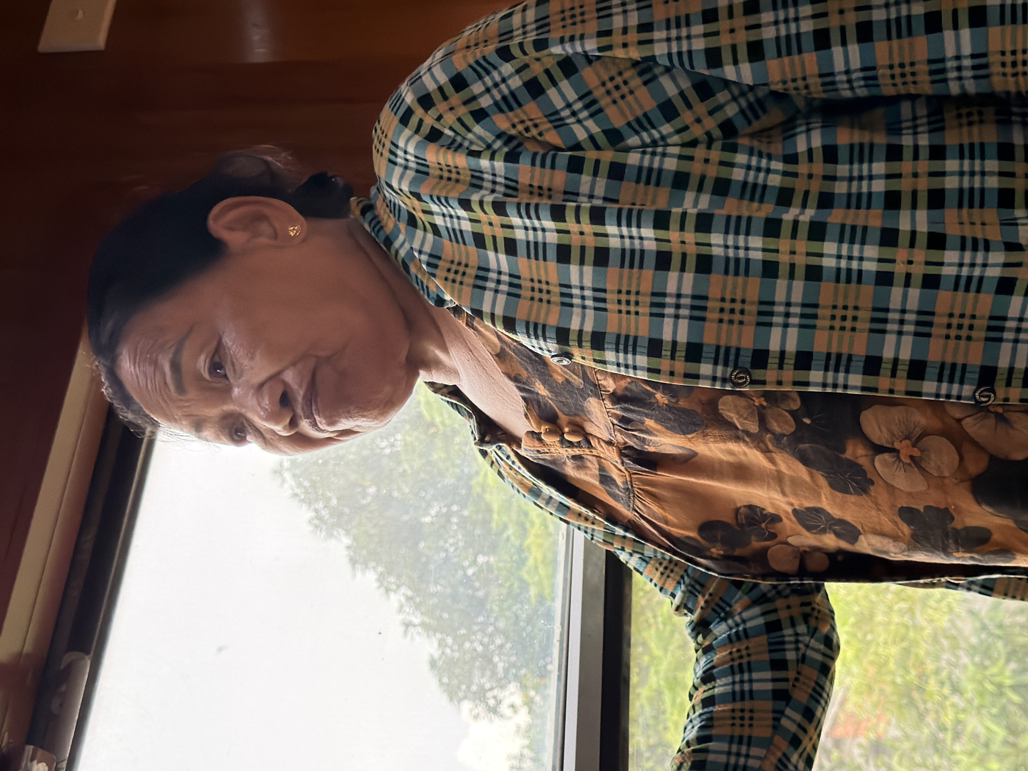 An elderly Asian woman is captured in a medium shot, standing indoors by a large window. She is looking out of the window, her expression thoughtful. She has dark hair pulled back and is wearing a patterned orange top underneath a plaid shirt. The plaid shirt features a pattern of teal, yellow, black, and white squares. The background outside the window is blurred, showing green foliage that suggests a natural, possibly park-like or rural setting, consistent with areas outside the immediate city center of Đà Nẵng, Vietnam. The interior setting appears to be a train carriage or a room with wooden paneling visible along the top and right edge of the frame.