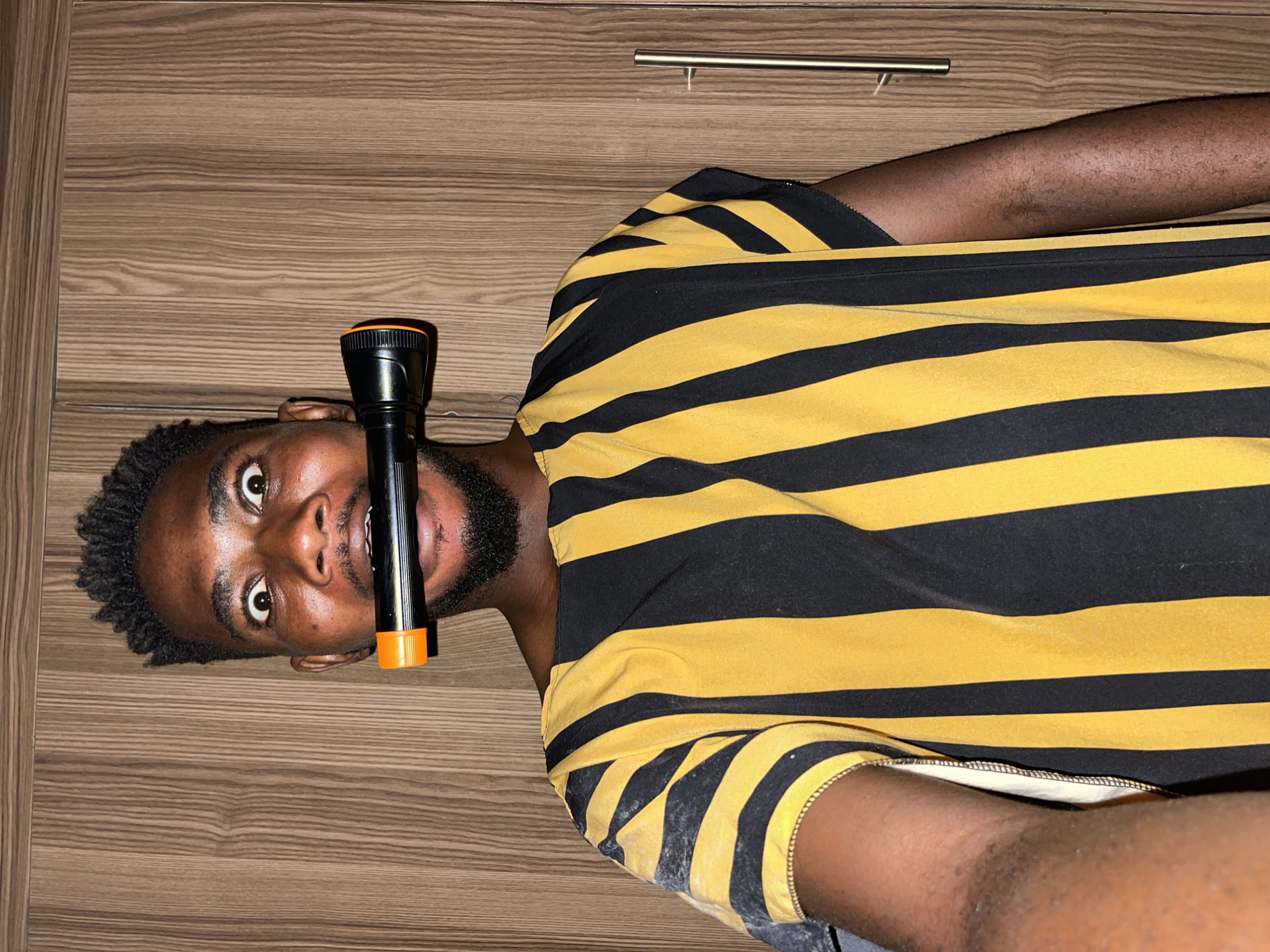 A young adult Black man is pictured from the chest up, lying on a light brown wooden surface, possibly a floor or large wall paneling, featuring a distinct wood grain pattern. The image is oriented such that he appears to be lying on his back, looking directly up at the camera. He has short, dark, curly hair and a beard/stubble on his chin and jawline.
He is wearing a short-sleeved t-shirt with bold horizontal black and yellow stripes. A black and orange flashlight is held horizontally in his mouth, resting just above his chin. His eyes are wide and expressive, conveying a look of surprise, amusement, or playful intensity.
The background includes a golden-colored metal handle, possibly from a drawer or cabinet, indicating an indoor setting. The lighting is even, suggesting artificial illumination rather than direct sunlight. There is no visible text in the image. The scene takes place in Muchiya, Nigeria.
